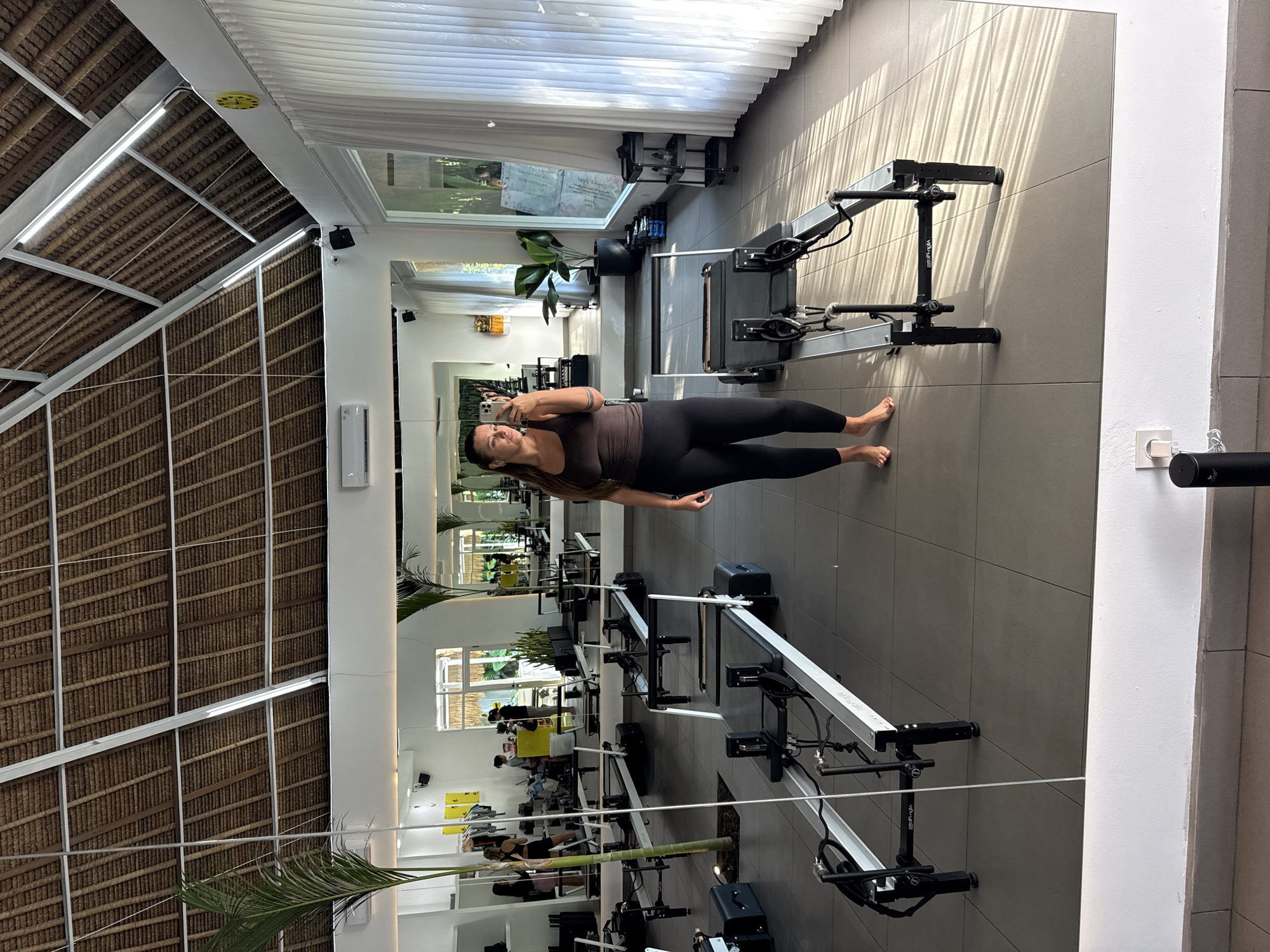 A woman wearing a brown top and black leggings stands in a gym, taking a selfie in front of a mirrored wall, with rows of pilates reformer machines in the background. The gym features a high thatched roof and windows offering natural light. The location is in Ubud District, Indonesia.