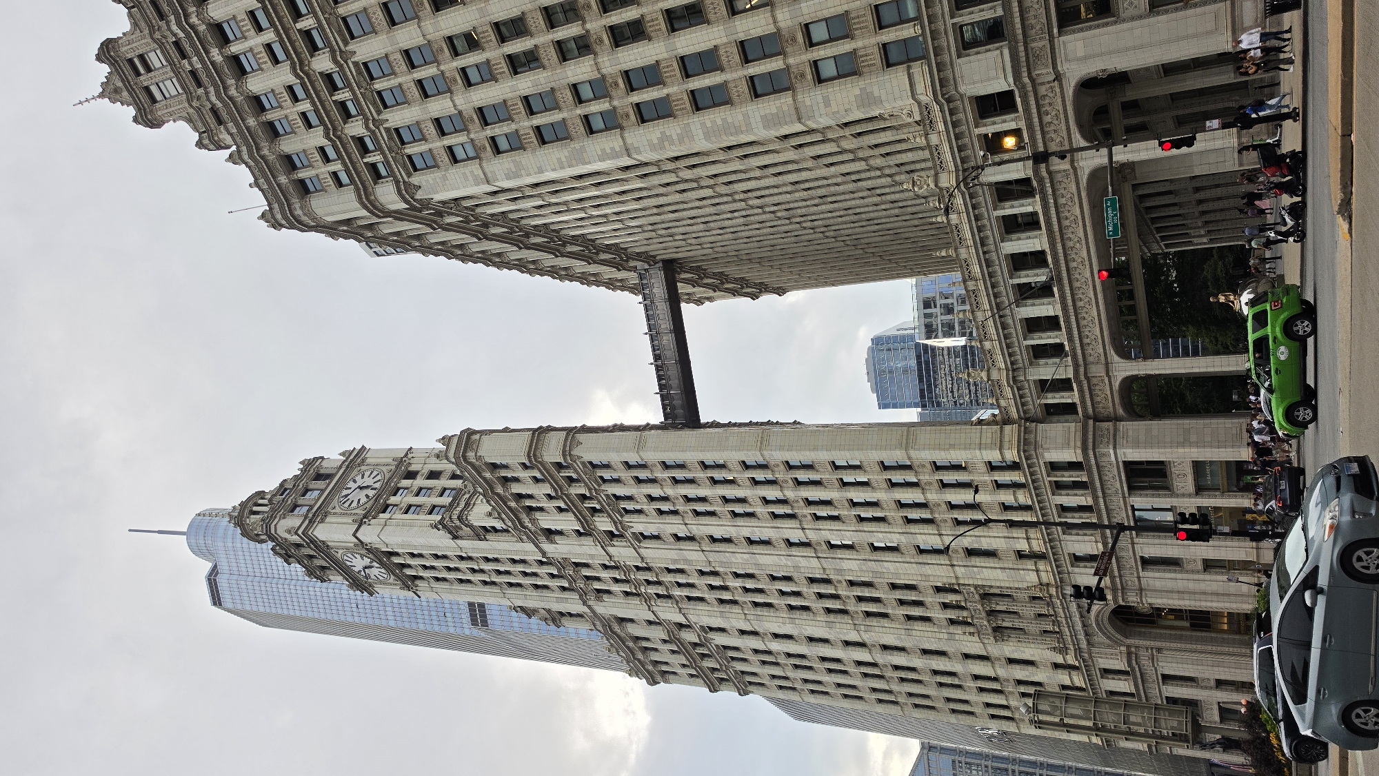 This is a photograph of the historic Wrigley Building and a neighboring building in Chicago, Illinois, United States of America, as seen from street level. The Wrigley Building, with its prominent clock tower, is on the left, and the Tribune Tower, another iconic Chicago skyscraper, is on the right. Both buildings are constructed of limestone and feature intricate architectural detailing. A skybridge connects the two buildings high above the street.
In the foreground, a silver Toyota Prius is driving through an intersection with a green light. A bright green SUV is also visible in the intersection. Pedestrians are walking on the sidewalks. Traffic signals are visible, indicating normal city traffic flow. The sky is overcast, suggesting a cloudy day. The overall impression is of a bustling urban environment with significant historical architecture.
