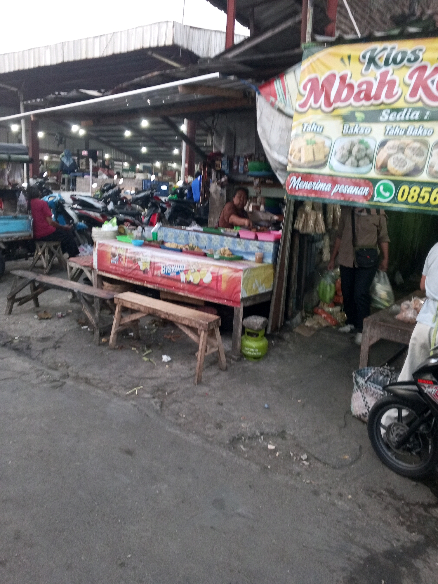 The image depicts an outdoor market scene, likely in Gentan, Indonesia, based on the provided context. A stall with a yellow and red banner advertising "Kios Mbah K" is prominently displayed, featuring images of "Tahu Bakso" and "Tahu" (tofu and meatballs). The banner also includes a WhatsApp number, "0856".
To the left of the banner, a person wearing a red shirt and dark pants sits on a stool next to a blue cart. In the background, numerous motorcycles are parked under a covered structure with visible lighting. A woman in a brown shirt stands behind a food stall. Wooden benches are placed in front of the food stalls. A green gas cylinder is visible on the ground near one of the stalls. The ground surface is asphalt, with some debris scattered around. To the right, a person in a light-colored shirt is partially visible, and a motorcycle's wheel and part of its body are in the foreground. The overall environment appears to be a bustling marketplace.