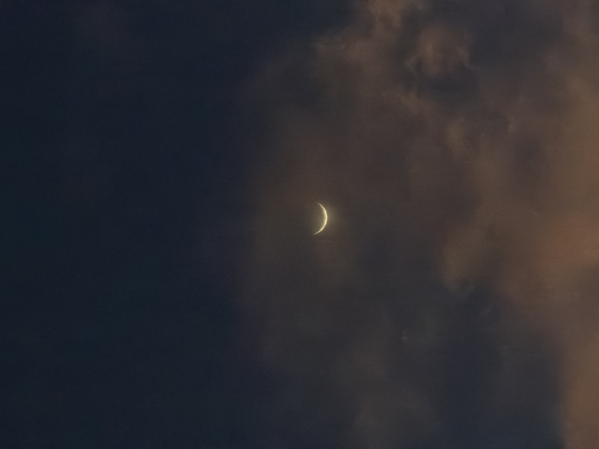 The image depicts a crescent moon in the night sky. The sky is partially obscured by dark clouds, with some areas appearing brownish-orange, possibly due to light pollution or sunset remnants. The location, Mengwi, Indonesia, is only relevant as geographical context, not visibly represented in the image.