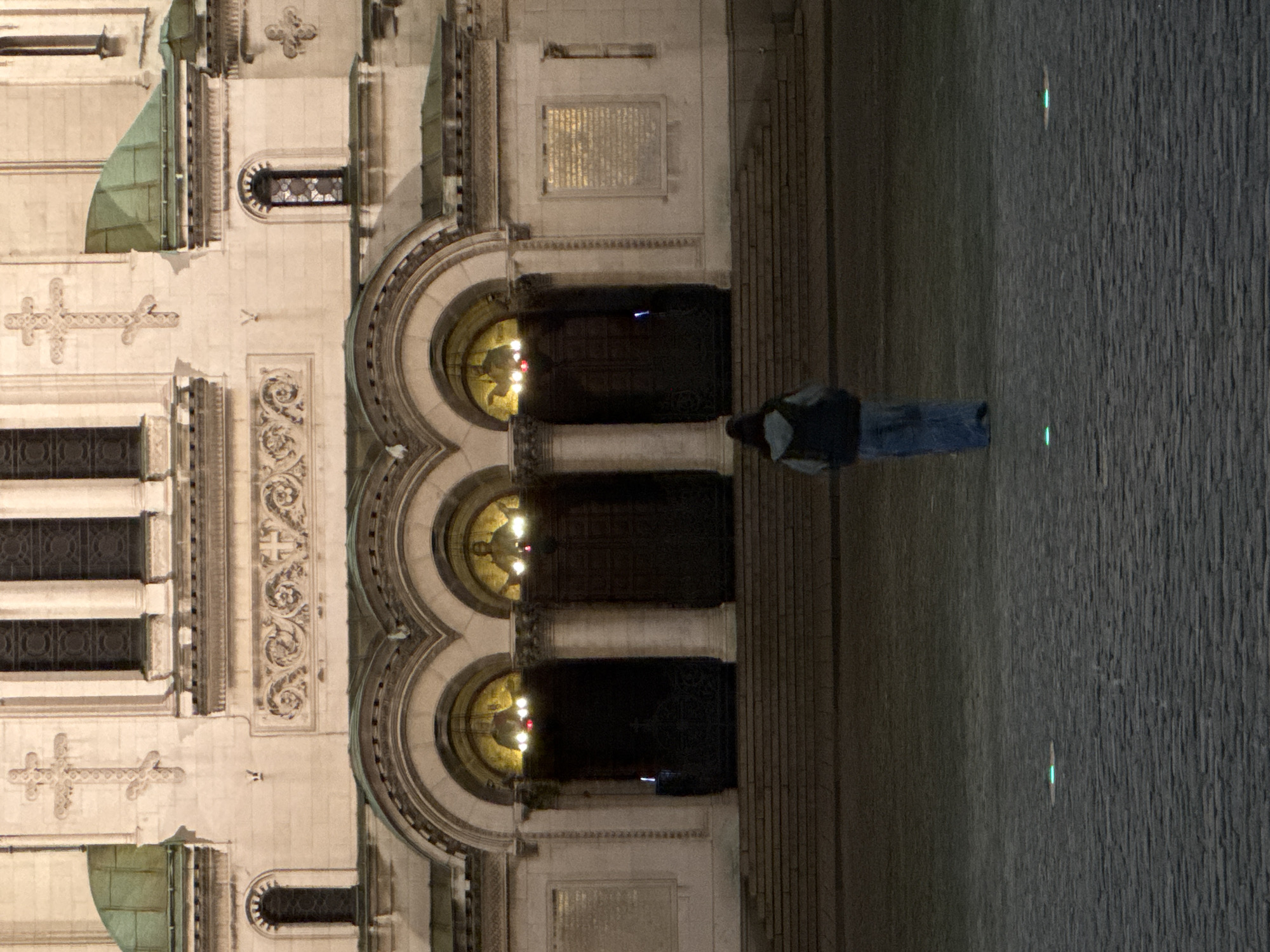 The image shows the Alexander Nevsky Cathedral in Sofia, Bulgaria, at night. The cathedral's facade is illuminated, revealing intricate architectural details, including crosses, decorative carvings, and three arched doorways. Inside the doorways, lights are visible. A person stands on the paved area in front of the cathedral, facing the entrance. Two small green lights are visible on the paved ground.