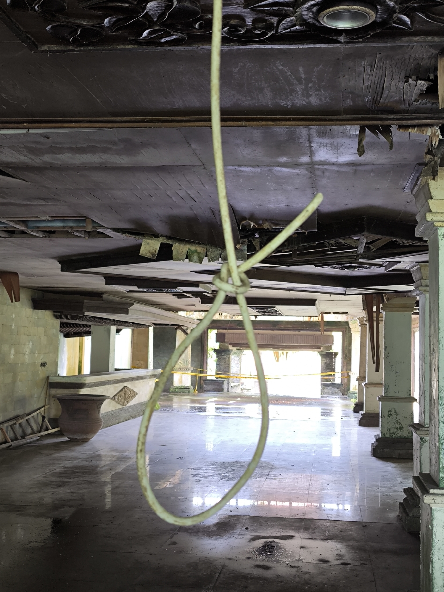 The image depicts an abandoned interior space with a noose hanging from the damaged ceiling. The location appears to be a building in disrepair, possibly in Mekarsari, Indonesia, as specified.
**Environment:**
The building shows significant decay. The ceiling has holes, exposing the structure above. The floor is reflective, possibly due to water accumulation. Columns are present, showing signs of aging and deterioration. There is also a ladder leaning against a counter, and yellow caution tape is visible in the background.
**Object:**
A noose hangs prominently in the center of the frame.
**People/Subjects:**
There are no people visible in the image.
