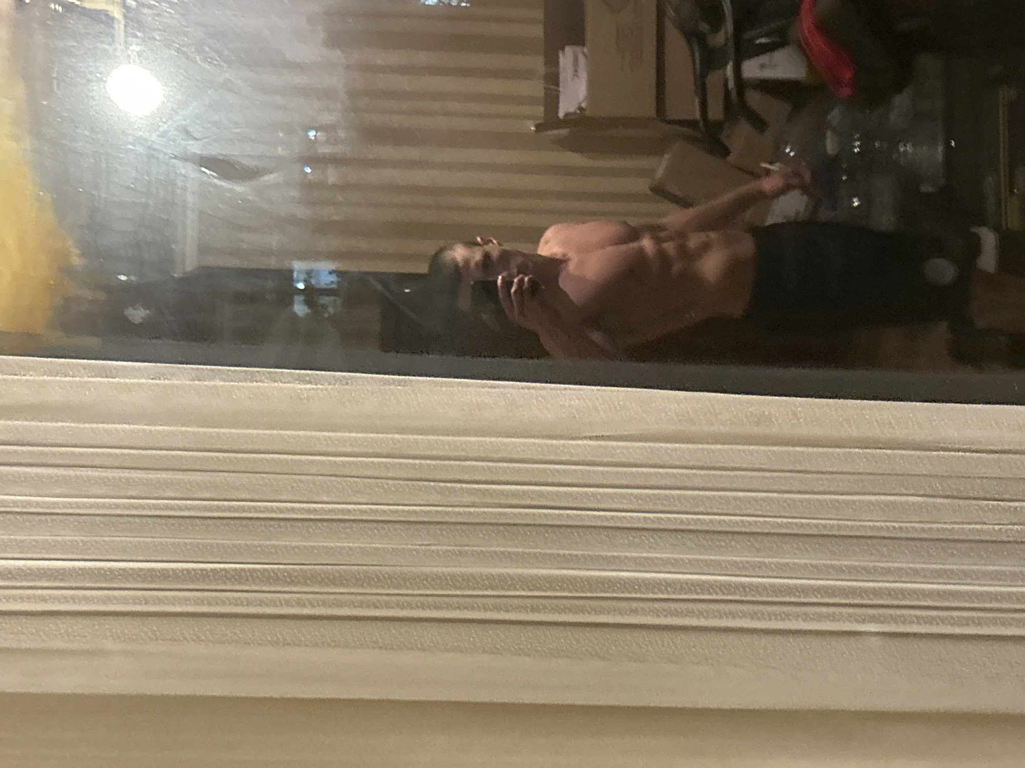 The image shows a shirtless man with dark hair and a muscular physique lying down indoors, possibly on a bed or floor. He is wearing dark shorts and appears to be holding or talking on a cell phone. The scene is observed through a window with closed Venetian blinds in the foreground. The reflection in the window shows an interior setting with office furniture. The city and country are not determinable from the image.
