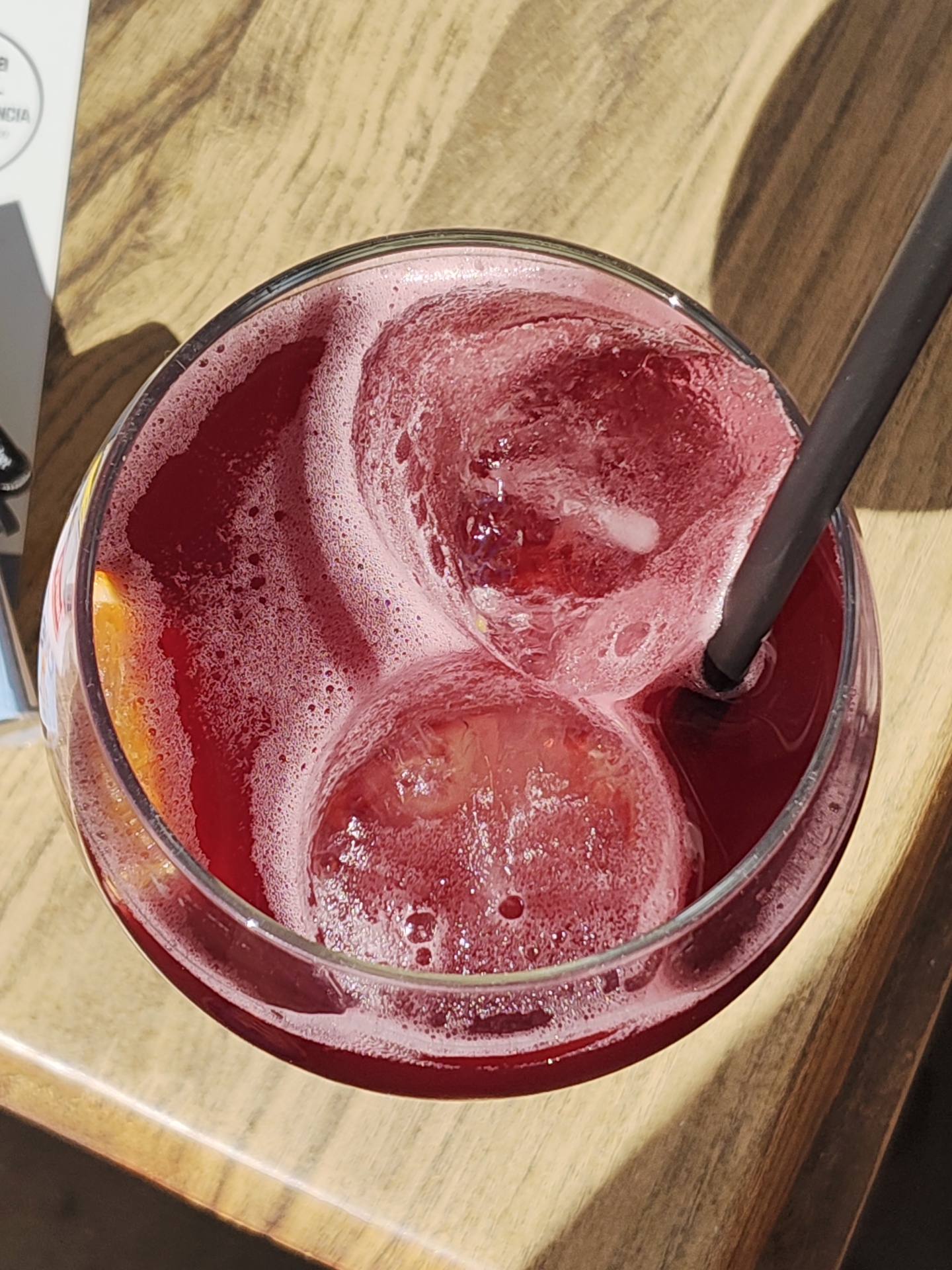 View from above of a red alcoholic drink (probably sangria) in a glass with ice cubes and a black straw, served outdoors on a wooden table. A slice of orange is visible inside the glass. The scene is likely in Dénia, Spain, though not confirmed by explicit elements in the image.
