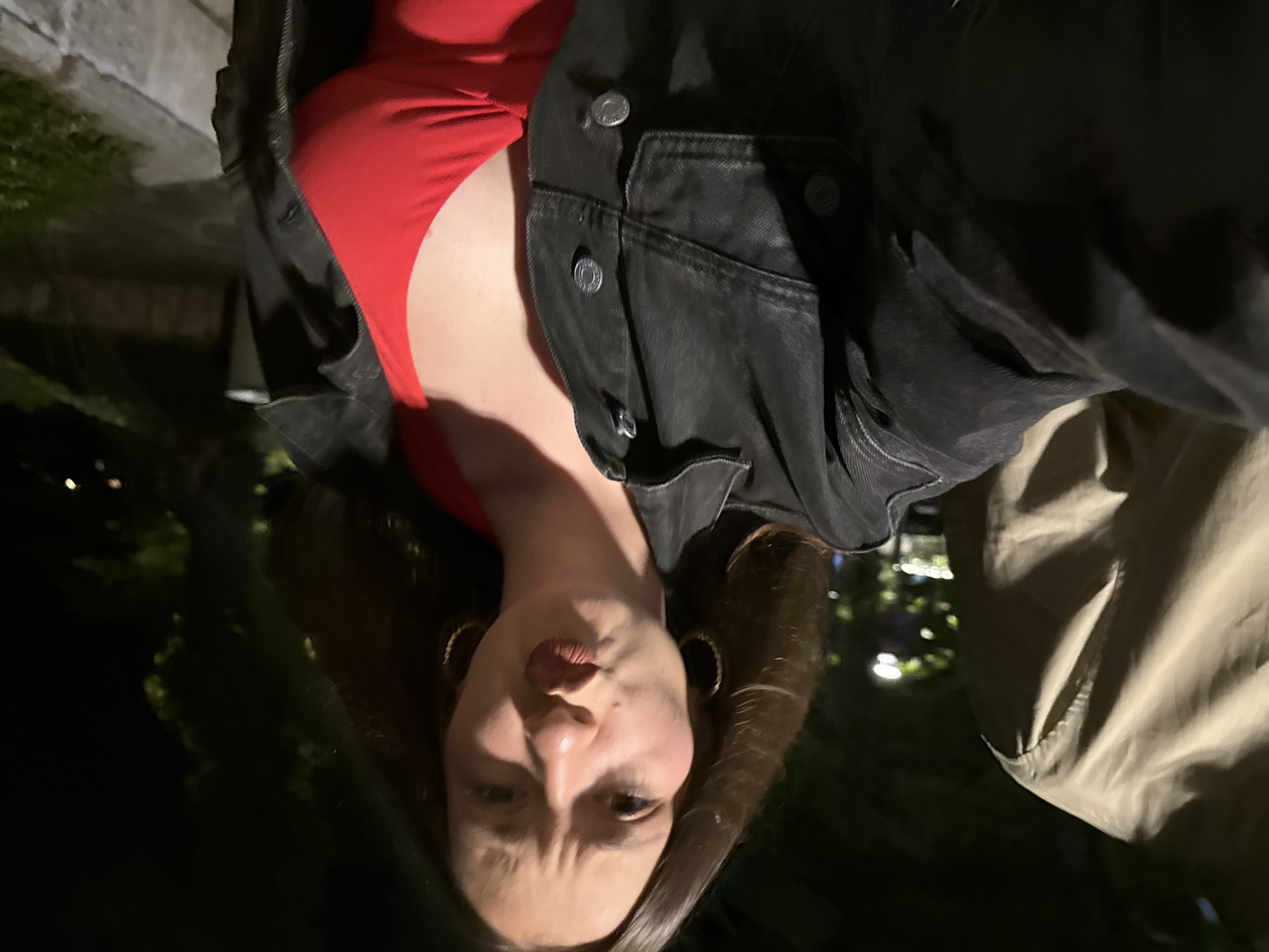The image depicts a nighttime outdoor scene in Sozopol, Bulgaria, captured from a low-angle perspective, likely a selfie. A female subject occupies the central and right portions of the frame, looking directly at the camera with a puckered lip expression and slightly furrowed brows. She has dark brown, shoulder-length hair and wears gold-colored hoop earrings. Her attire consists of a red V-neck garment, over which an open, dark-colored denim jacket with metallic buttons is worn. The jacket's pockets and placket are visible. To the far left, the shoulder and upper arm of a second individual, clad in a light-colored (beige or light brown) garment, are partially visible. The background is dark, featuring blurred green foliage and indistinct points of artificial light, indicative of streetlights or distant building illumination. A low concrete or stone barrier extends along the bottom right edge of the frame. The overall lighting is dim, consistent with a nocturnal setting.