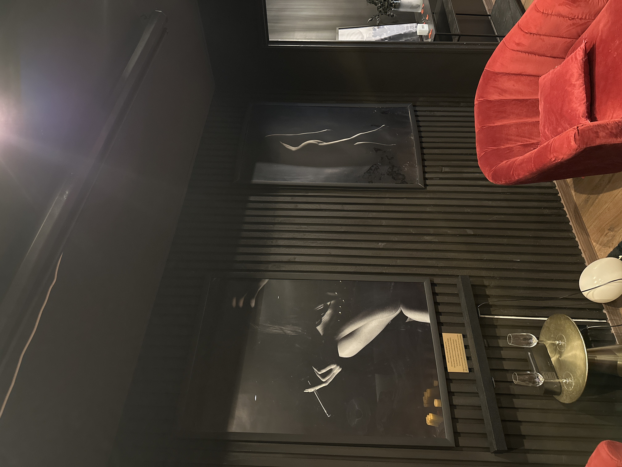 The image depicts an indoor scene with a dimly lit, moody ambiance. The walls are covered in dark, vertical paneling. Two large framed black and white photographs are prominently displayed on the wall. The photograph on the left features a woman holding a cigarette, and the one on the right showcases the silhouette of a nude figure. In the foreground, a plush red armchair with a matching cushion is positioned. To the left of the armchair, a small round metallic table holds two champagne flutes and a speaker. A white spherical lamp rests on the floor near the armchair. The overall setting appears to be a private room or lounge, potentially a bar or a residence, in Le Havre, France.