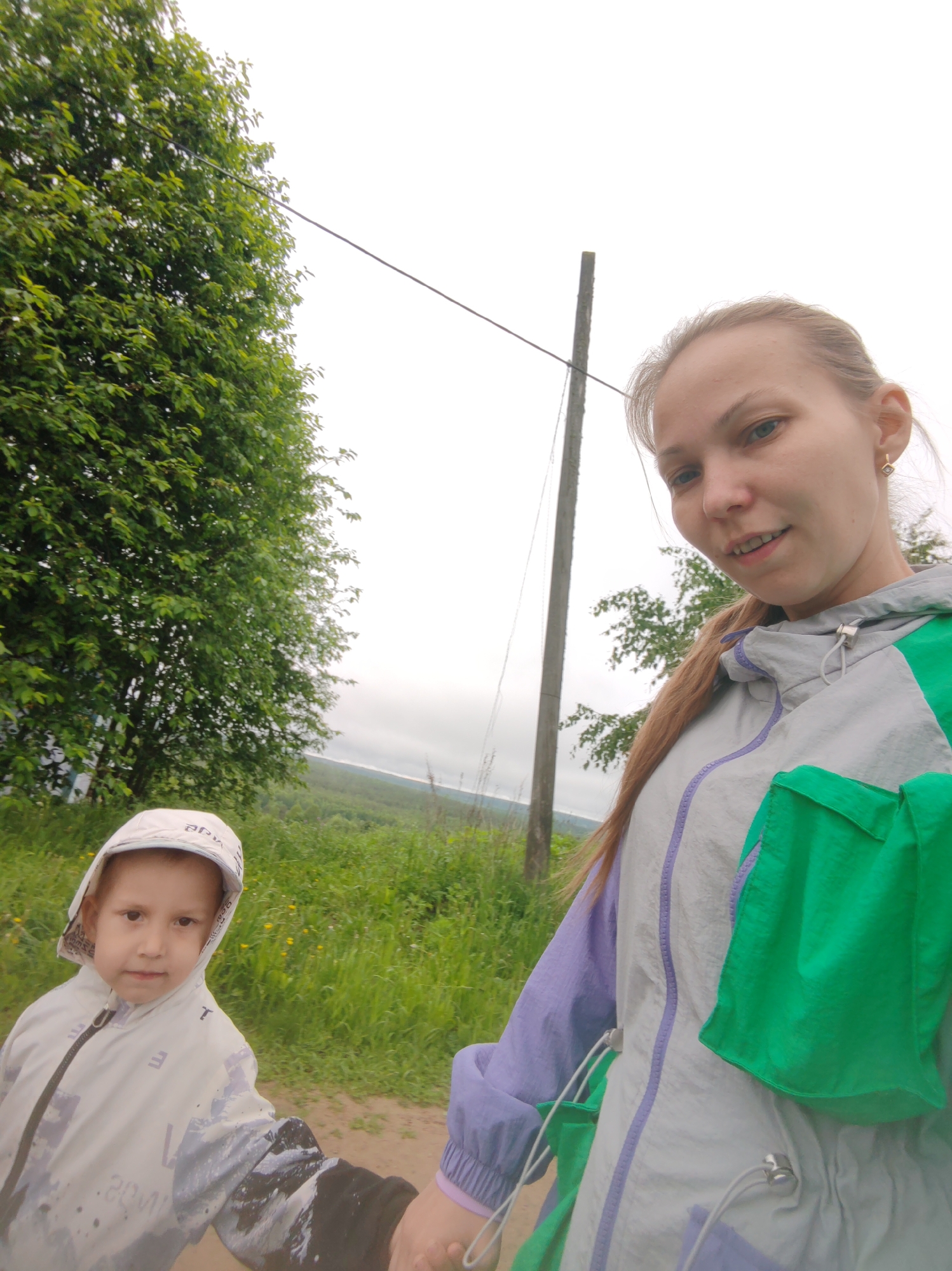 The image depicts a woman and a young child outdoors. The woman is on the right side of the frame, wearing a jacket with gray, green, and lavender panels. Her hair is pulled back, and she is looking at the camera. The child is on the left, wearing a hooded jacket with a muted, patterned design. They are holding hands, suggesting a relationship.
Behind them, there is a grassy field and a view of what appears to be a landscape with trees and possibly a body of water in the distance under an overcast sky. To the left of the woman, a wooden post is visible, possibly a utility pole.
The image seems to be taken in a rural or semi-rural location. The location is Чухлэм, Russia.