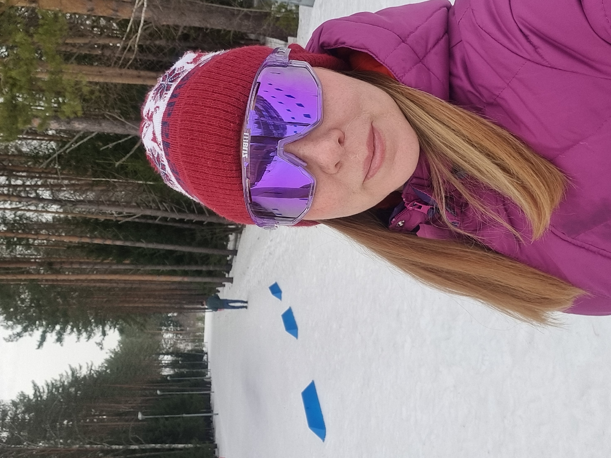The image displays a snowy outdoor scene in Прикамский, Russia. A female individual occupies the foreground, positioned in a selfie orientation, directly facing the camera. She wears a red knitted beanie with white and dark patterns, large purple mirrored sports goggles with the "BLIZ" brand visible, and a magenta quilted winter jacket with an orange interior lining. Her light brown hair is visible below the beanie and around her shoulders. In the background, a wide, snow-covered path extends into the distance, flanked on both sides by dense rows of tall pine trees. Several dark, slender poles, likely streetlights, are visible along the left side of the path. Three blue, wedge-shaped markers are placed on the snow-covered ground to the left of the path, receding in size towards the background. A male figure, wearing a dark green jacket and blue pants, walks away from the camera in the middle distance on the path. The sky is overcast.