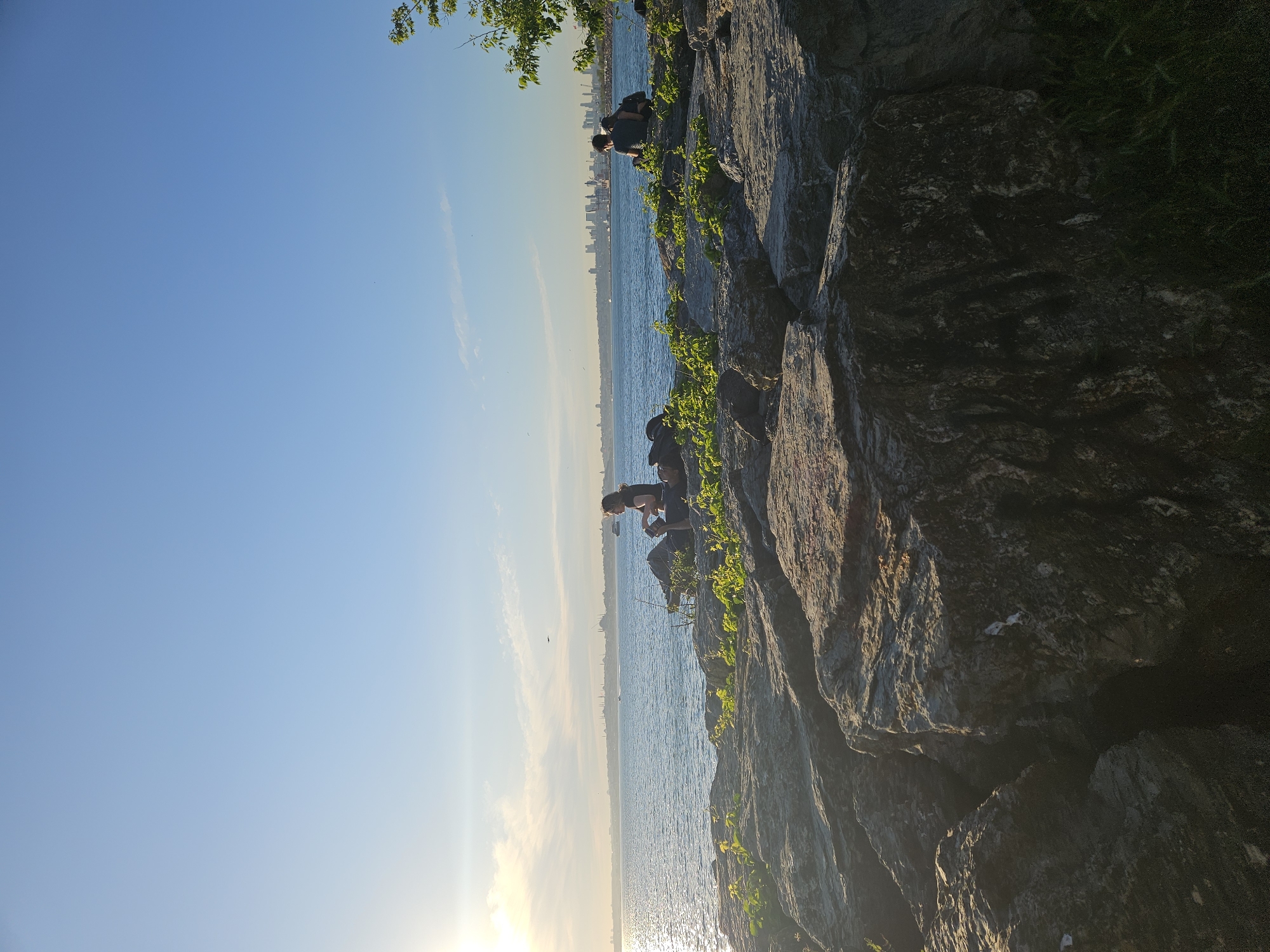 The image depicts a rocky coastline in Kadıköy, Turkey, during daylight. Two individuals are seen sitting and crouching on the rocks overlooking the water. In the distance, a city skyline is visible across the water. The sky is blue with some cloud cover, and the sun is low, casting a glare on the water. Greenery grows on the rocks along the shoreline.