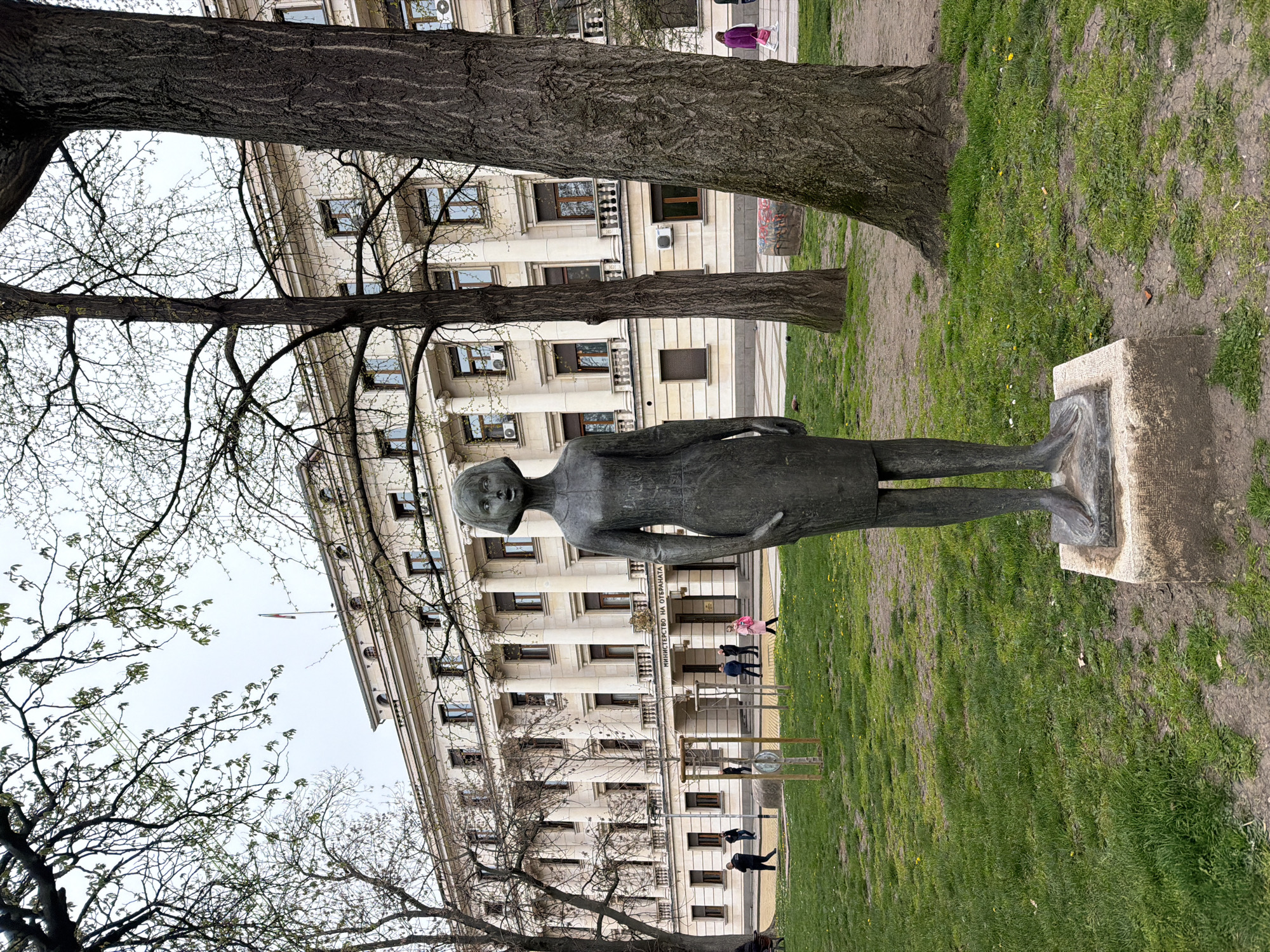The image shows a bronze statue of a young woman standing on a stone pedestal in a park-like setting. The statue is thin and elongated, with a simple dress. In the background is a large, ornate building with classical architectural features, identified as the Ministry of Culture in Sofia, Bulgaria. Several trees are present, one partially obscuring the view of the building. A few people are walking in front of the building. The grass appears patchy. The sky is overcast.