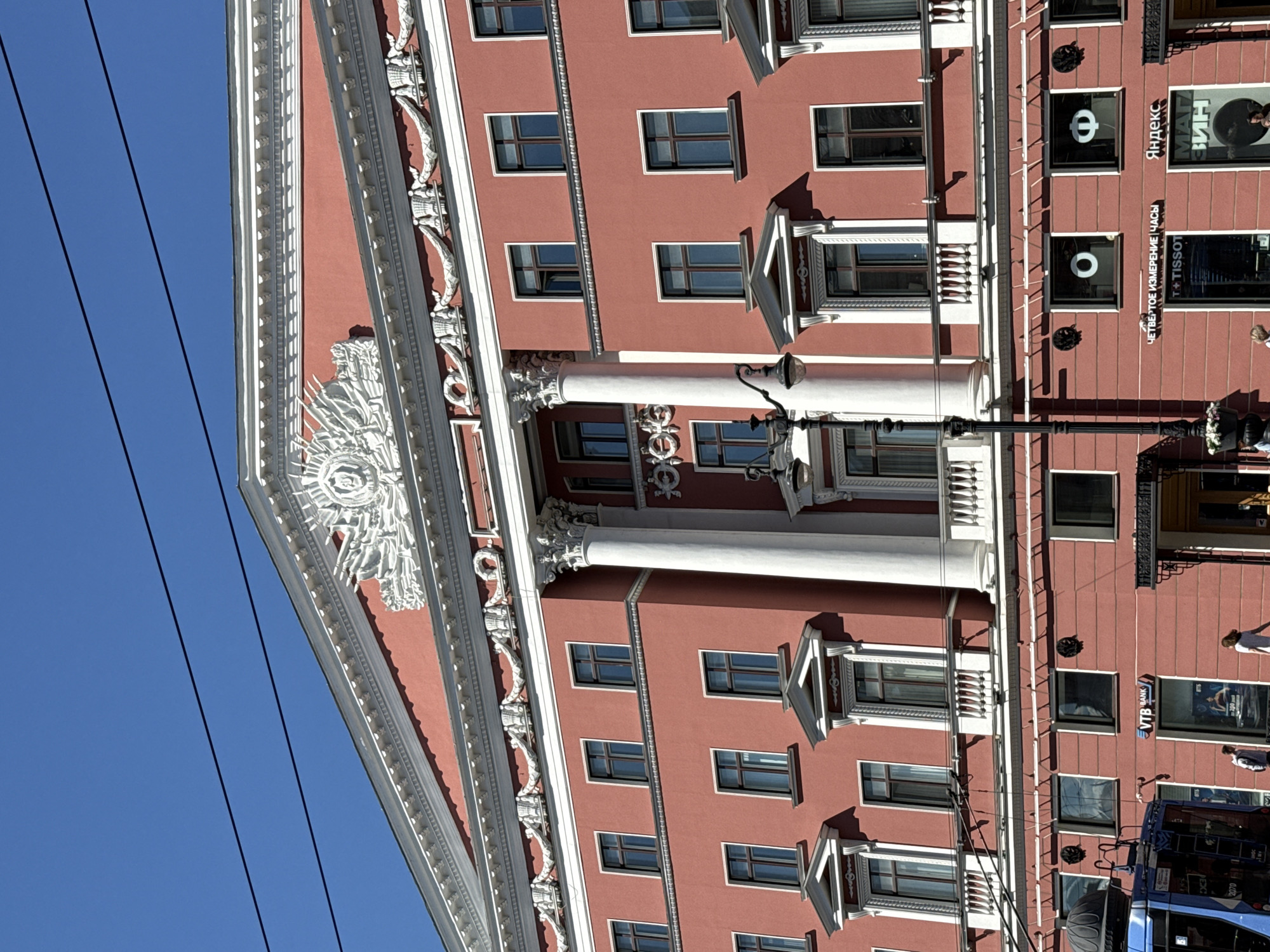 This image shows the exterior of a building with a red facade and intricate white architectural details. The building has multiple floors with windows and balconies. In the foreground, there are power lines against a clear blue sky.
Visible text includes "Яндекс" (Yandex), "ЧЕТВЕРТОЕ ИЗМЕРЕНИЕ ЧАСЫ" (Fourth Dimension Watches), "T+TISSOT", and "VTB BANK". There are also logos that appear to be for Yandex and Tissot.
Several people are visible on the sidewalk in front of the building, suggesting it is a public space. One person is walking away from the camera, and another is standing near a bus. A blue bus is partially visible on the right side of the image. The lighting indicates it is daytime, likely sunny, casting shadows on the building. The overall scene appears to be a street view of a commercial area in Saint Petersburg, Russia.