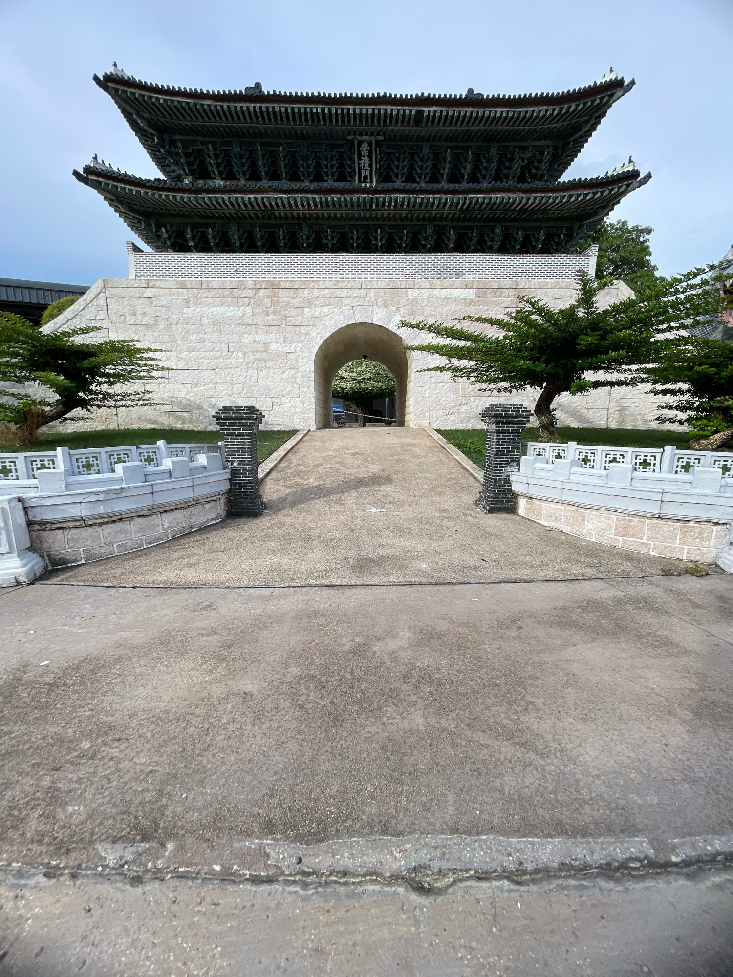 A wide-angle outdoor shot captures a traditional Korean-style gate structure, which appears to be a miniature replica, located in Pattaya City, Thailand. The gate is the focal point, with its dark, multi-tiered roof featuring intricate green detailing and upturned eaves. Beneath the roof is a section of white brickwork, leading to a large arched entryway. The entrance is framed by a pathway of concrete, flanked by decorative white stone railings on either side. Small, manicured trees with dark green foliage are positioned on either side of the pathway, partially obscuring the white brick wall. The ground in the foreground is a concrete pathway with some texture and discoloration. The sky above is mostly clear with a few wispy clouds. There are no people or animals visible in the frame. The overall impression is of a static, architectural display.