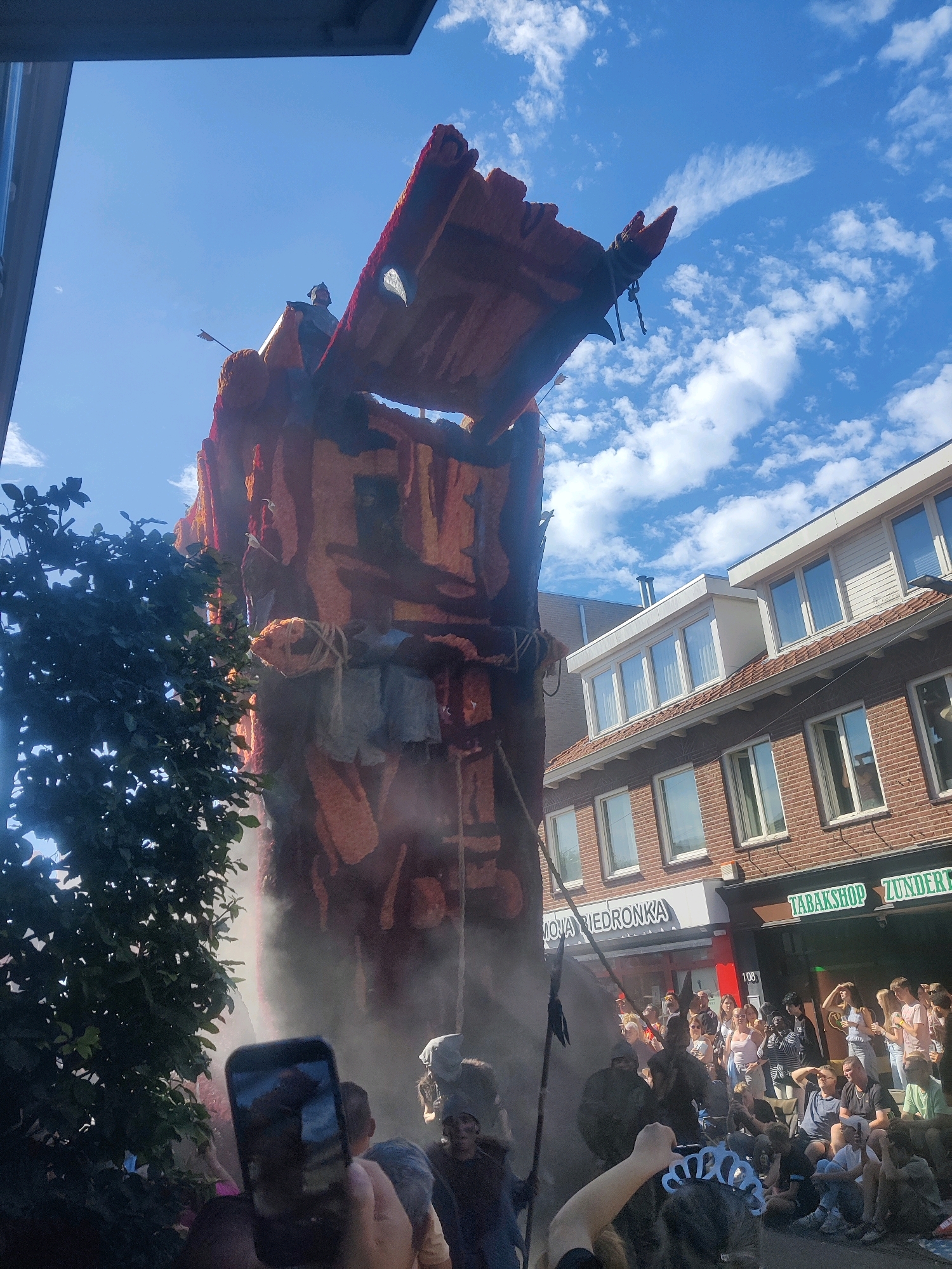 The image depicts a scene in Zundert, Netherlands, during what appears to be a parade or festival. In the foreground, a large, elaborately decorated float resembling a tower or structure is prominent. The float is adorned with a rough, textured material in shades of orange and red, and several figures appear to be part of its construction or decoration, including what looks like a figure in armor with an arrow protruding from it. Smoke or mist emanates from the base of the float.
A crowd of onlookers is gathered on the street, watching the float pass by. Many are holding up their phones to take pictures or videos. There are also individuals dressed in costumes, including one person with a tiara. In the background, buildings line the street, some of which have businesses with signage visible, including "MOJA BIEDRONKA" and "TABAKSHOP ZUNDER". The sky is blue with scattered white clouds. The overall atmosphere suggests a lively public event.