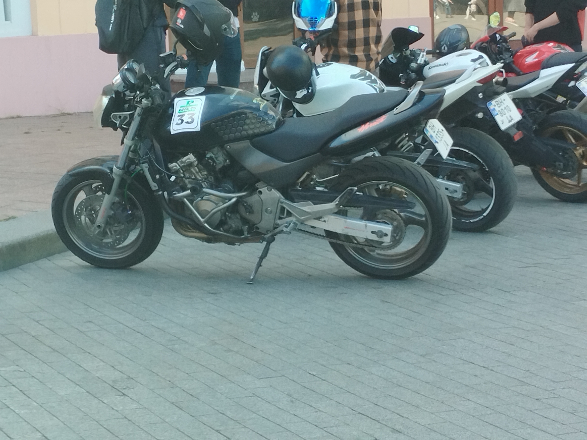 The image shows three parked motorcycles on a paved surface in what appears to be an urban setting. The first motorcycle is a black standard motorcycle with a number "33" displayed on its fuel tank. The second motorcycle is white, and the third is a red and white sport bike. People are standing behind the motorcycles. The location is Odesa, Ukraine.