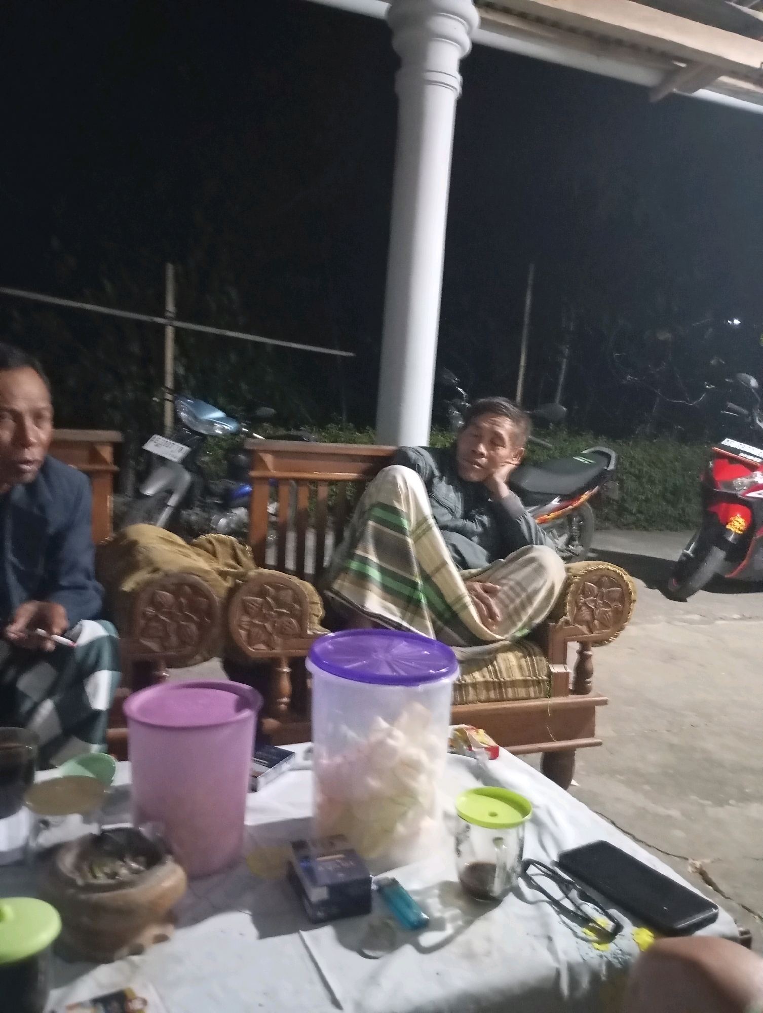 The image depicts an outdoor scene at night, possibly in Wonogondo, Indonesia. In the foreground, a white table is covered with a white cloth. On the table are several objects, including a purple-lidded container, a clear plastic container with a purple lid filled with what appears to be snacks, a small glass with a green lid containing a dark liquid, a lighter, a pair of dark-rimmed glasses, and a black smartphone. An ashtray with cigarette butts is also visible. To the left, partially visible, is a person wearing a dark jacket and a patterned shirt. To the right and slightly further back, a man is seated on a wooden chair with a woven seat and back. He is wearing a dark jacket and light-colored patterned pants, and has his hand resting on his cheek, appearing relaxed or contemplative. Behind the seated man, a motorcycle is parked. Further in the background, another motorcycle is visible. The environment appears to be a residential or communal outdoor area, with a white pillar visible on the left, supporting an overhang. The lighting suggests it is nighttime.