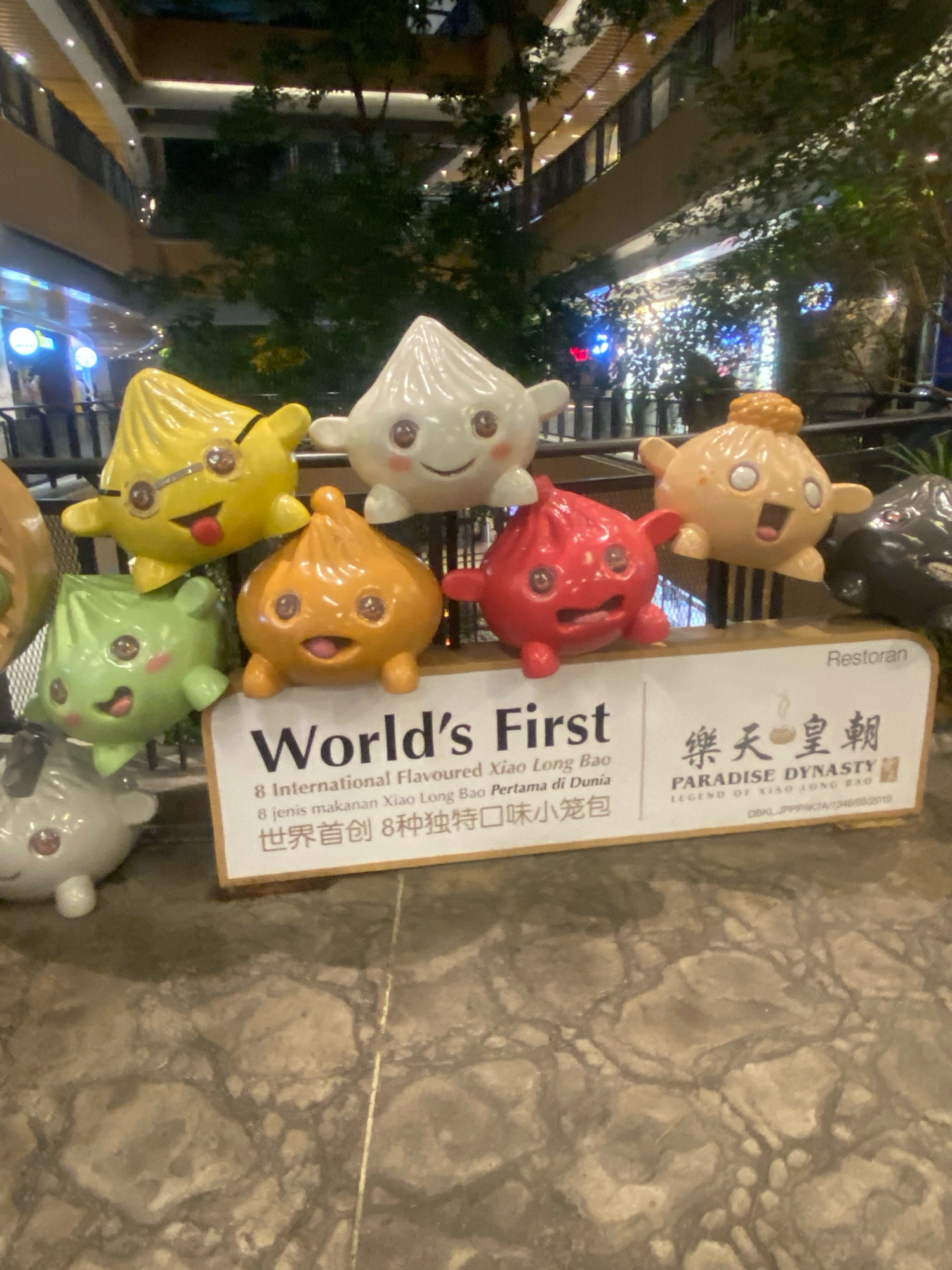 In Kuala Lumpur, Malaysia, at the Paradise Dynasty restaurant, eight animated dumpling characters are displayed on a white base. The dumplings represent eight international flavors of Xiao Long Bao. Each dumpling has a unique color and facial expression, with some wearing glasses or holding their hands up. The backdrop features the restaurant's logo in Chinese and English, along with the phrase "LEGEND OF XIAO LONG BAO." The scene is set in an indoor dining area with a marble floor and a blurred background of trees and building structures, suggesting an evening setting.