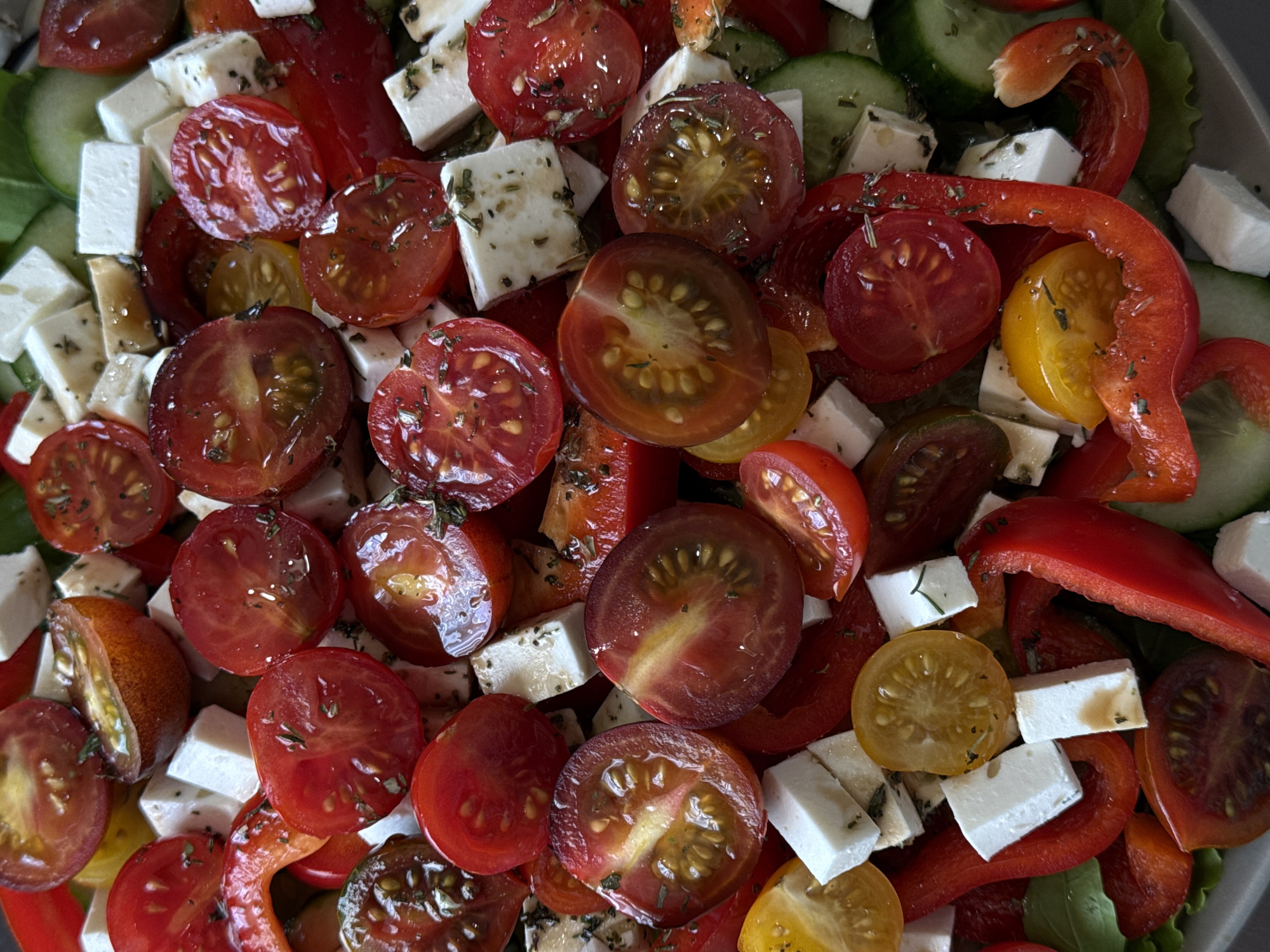 The image shows a close-up view of a salad. The salad consists of halved red and yellow cherry tomatoes, cubed feta cheese, sliced red bell peppers, and cucumber slices, all seasoned with herbs.