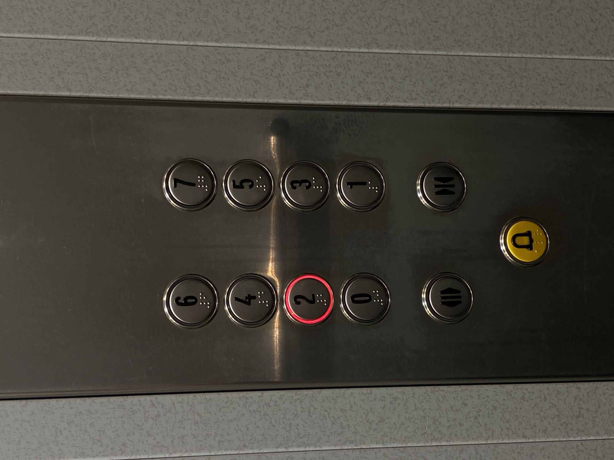 The image shows an elevator button panel with buttons arranged in two rows. The top row contains buttons for floors 7, 5, 3, 1, the door open symbol and a button that has the letter "D" on it, colored yellow. The bottom row contains buttons for floors 6, 4, 2, 0, and the door close symbol. The button for floor 2 is illuminated with a red light. The button panel is made of a brushed metal material and is set into a wall with a mottled gray finish. This is likely an elevator panel found somewhere in the city of Puebla, Mexico.