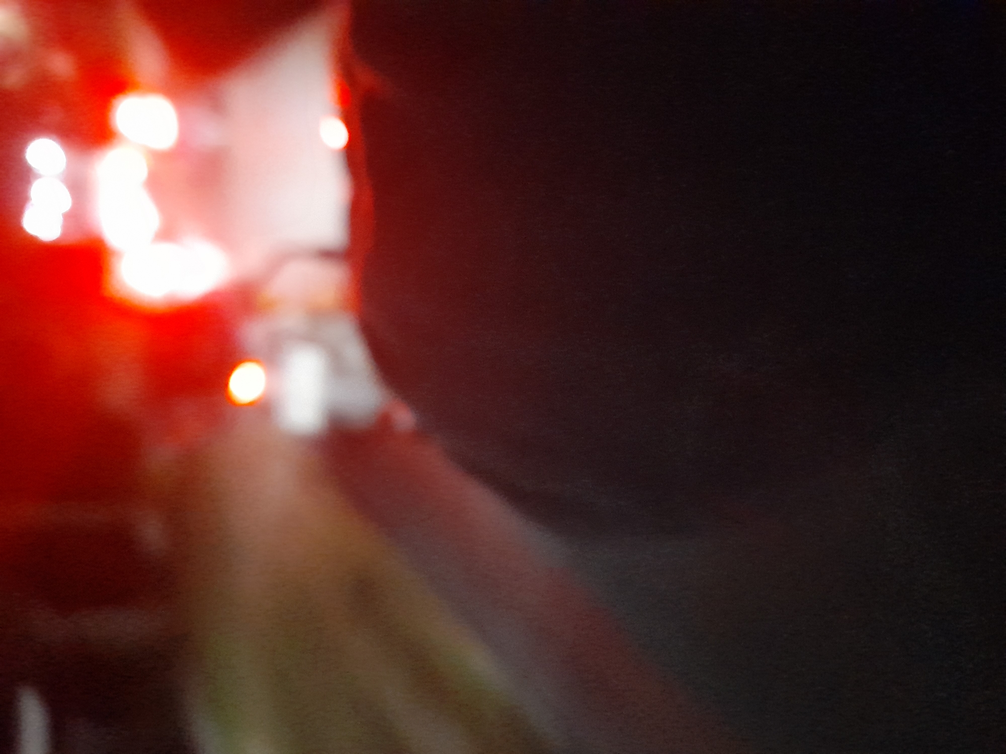 The image shows a blurred, out-of-focus scene with a prominent dark foreground that appears to be the back of someone's head and shoulders. In the background, there are bright, out-of-focus red and white lights, suggesting an urban environment at night, possibly with vehicle taillights or streetlights. The scene is undated and lacks specific identifying features to pinpoint its location within Jetaksari, Indonesia. There are no discernible people or specific objects in focus.