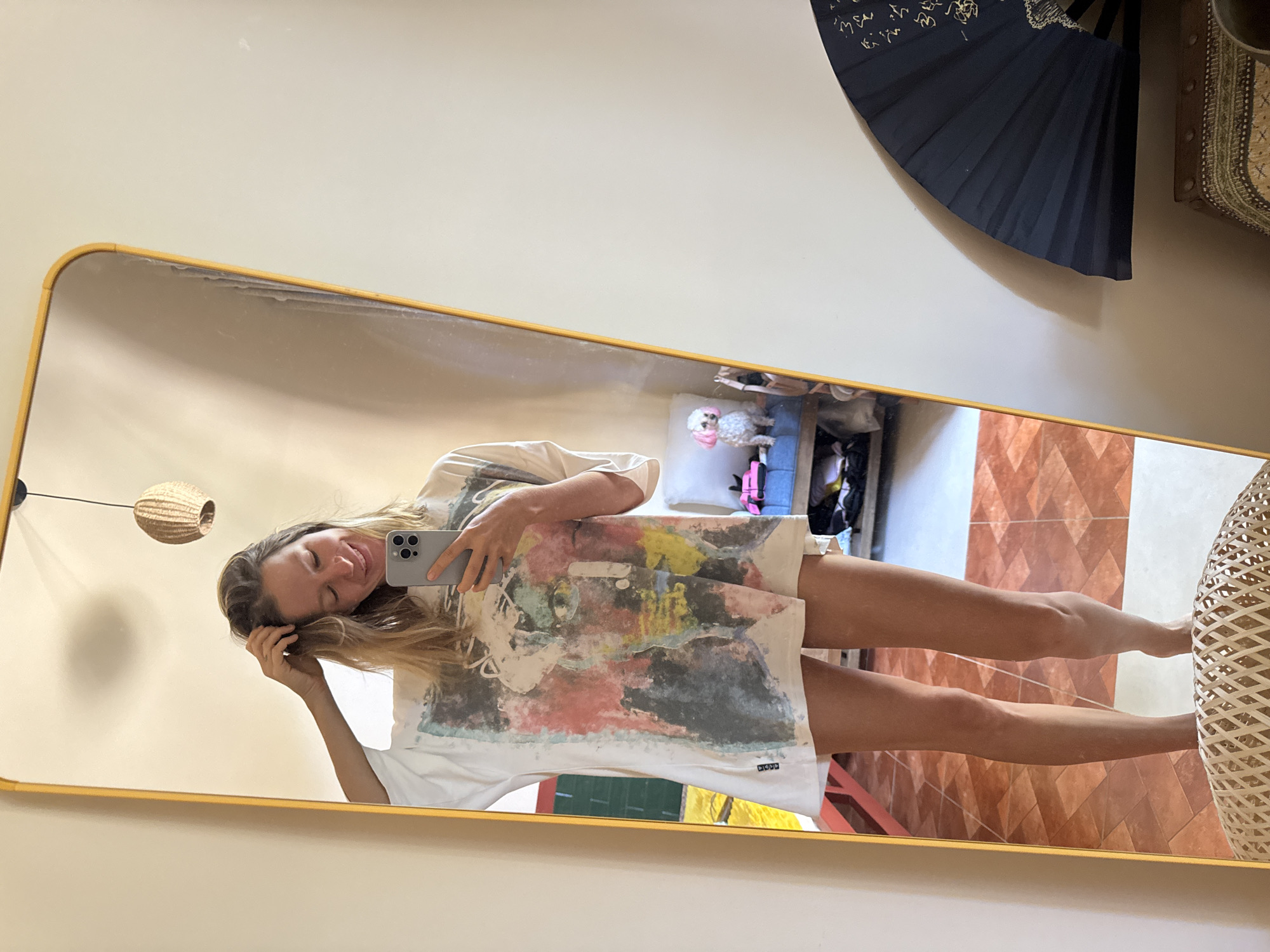 A full-body reflection shows a young woman in a large, graphic print t-shirt dress standing in what appears to be an indoor space. She is holding a smartphone in her right hand and her left hand is raised, touching her hair. The background of the reflection includes a floor with terracotta-colored tiles, some furniture, and a small white dog with pink accents on its ears. To the right of the image, a dark, ornate fan is partially visible. The overall impression is of a casual, personal moment captured in a home environment.