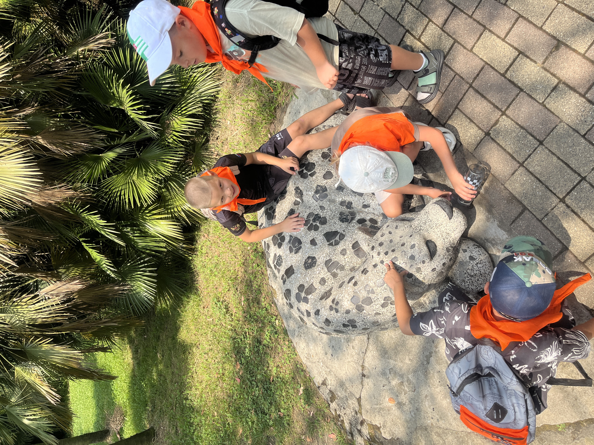 In Sochi, Russia, four children, three boys and one girl, are gathered around a large, grey, stone sculpture of an animal, possibly a leopard or jaguar, which is embedded in the ground. The sculpture has dark grey spots. One boy, wearing a black t-shirt and shorts, sits on the sculpture and looks at the camera. Another boy, wearing an orange shirt, shorts, and a white baseball cap, kneels beside the sculpture, holding a clear bottle with a black lid. To the right, a third boy in an orange bandana and a white t-shirt, stands looking down at the sculpture. The fourth child, a girl wearing an orange bandana and a blue and green baseball cap, is lying on the ground, reaching towards the sculpture. All four children are wearing orange bandanas around their necks. The sculpture is on a paved pathway, and behind them are lush green palm trees and grass.