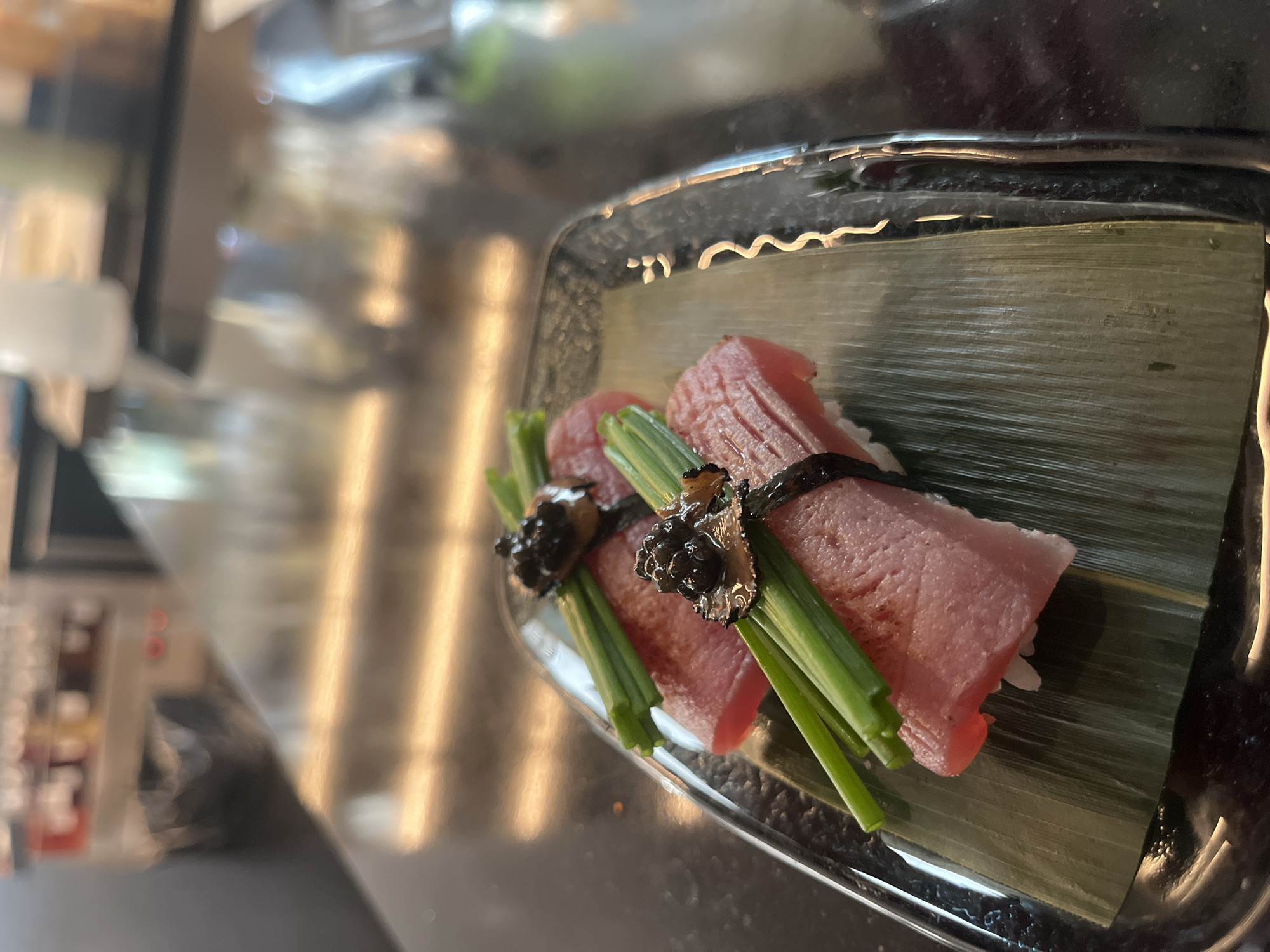 The image shows two pieces of what appears to be sushi or similar type of dish served on a rectangular plate lined with a green leaf. Each piece consists of a slice of raw fish (likely tuna) wrapped around a small bed of white rice. The fish is bound with dark strips, possibly seaweed, and garnished with green chives and black truffle shavings. The background is out of focus but appears to be a restaurant setting, potentially in the city of Sofia, Bulgaria.