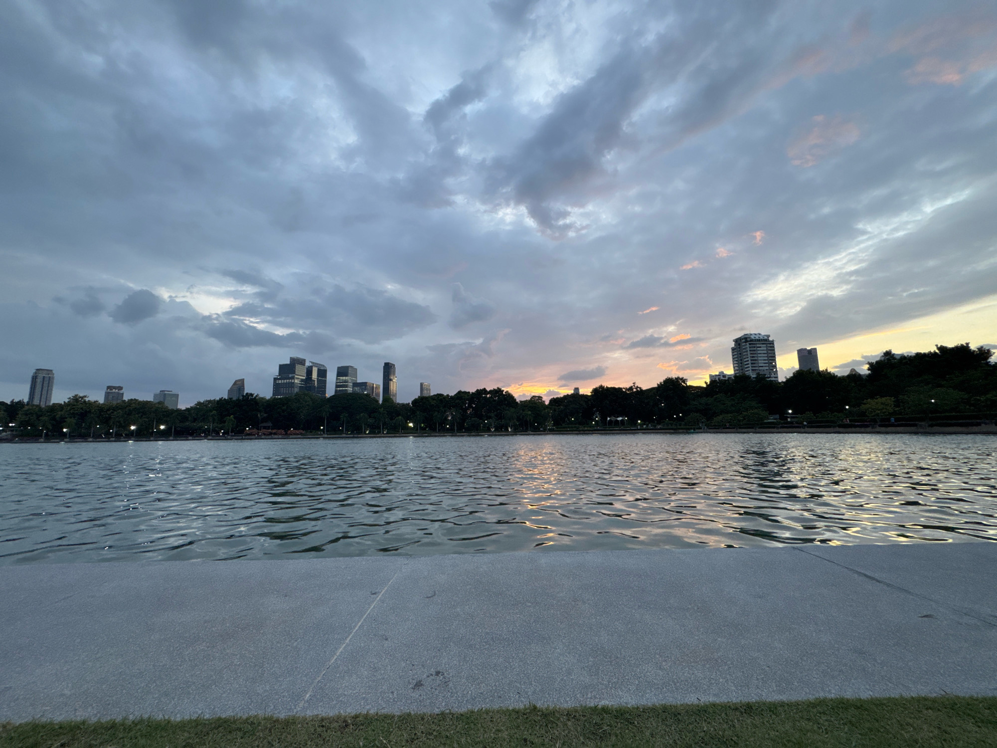 The image depicts a serene lake with gentle ripples reflecting the muted colors of a twilight sky. A paved walkway borders the foreground, with a strip of green grass beyond. In the background, a line of lush trees separates the lake from a cluster of modern skyscrapers, indicative of an urban setting. The sky is filled with dramatic clouds, some tinged with soft hues of orange and pink from the setting sun. The scene is likely set in Lumphini Park in Bangkok, Thailand, a popular urban oasis known for its central lake and surrounding cityscape. There are no people visible in the image, and the overall atmosphere is peaceful and tranquil.