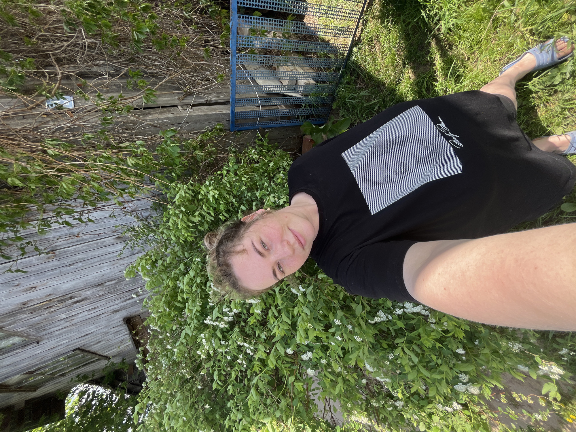 A woman is lying on a bush with white flowers in a yard in Сосново, Russia. She is wearing a black t-shirt with a white square graphic and signature on the front and blue sandals. A wooden building with a boarded window and a blue metal stairway are visible in the background, along with grass and trees.