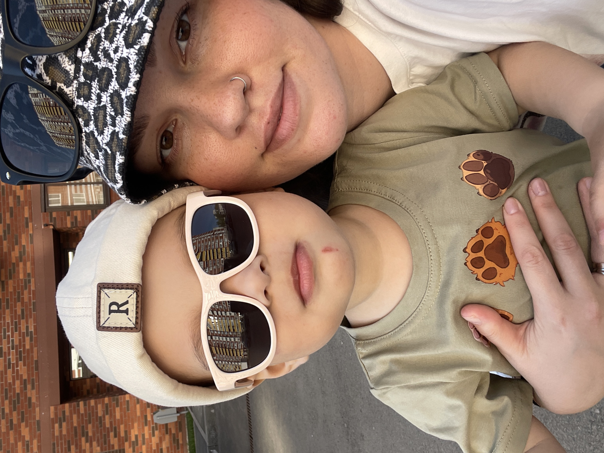 A woman and child pose for a photo outdoors in Izhevsk, Russia. The woman, with light skin and a nose ring, wears a patterned hat and sunglasses perched on top. The child, presumably lying on the ground with the woman leaning over, wears a light-colored cap with a "R" patch and light-framed sunglasses. The child's t-shirt is green and features paw print designs. The building reflected in the glasses indicates the location in Izhevsk, Russia. The surroundings appear to be a paved area adjacent to a brick building, with possible grass or vegetation visible.