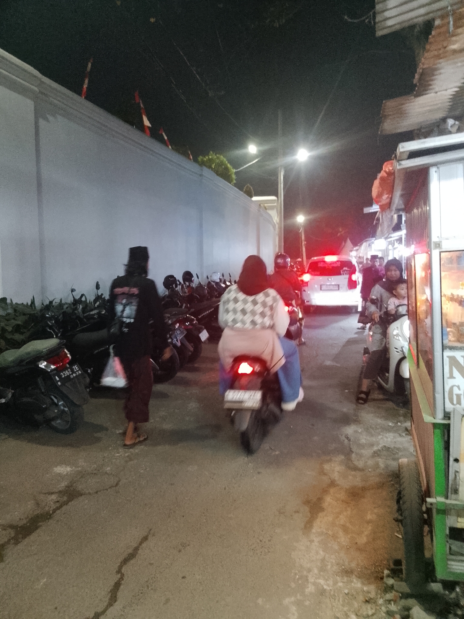 At night in Sukamantri, Indonesia, a street scene unfolds with several people and vehicles. A light-colored wall runs along the left side of the frame, with several red and white flags atop it. In the foreground, a person wearing a black long-sleeved shirt and dark pants walks away from the camera. To their right, multiple motorcycles are parked.
Further down the street, two people are riding a motorcycle, the passenger wearing a checkered headscarf and a light-colored jacket. A white car with its brake lights illuminated is in motion ahead of them. On the right side of the image, a stall with a corrugated metal roof is visible. A woman and a child are on another motorcycle to the right of the street. The ground is paved with asphalt and shows some signs of wear. Streetlights are present, casting light on the scene.