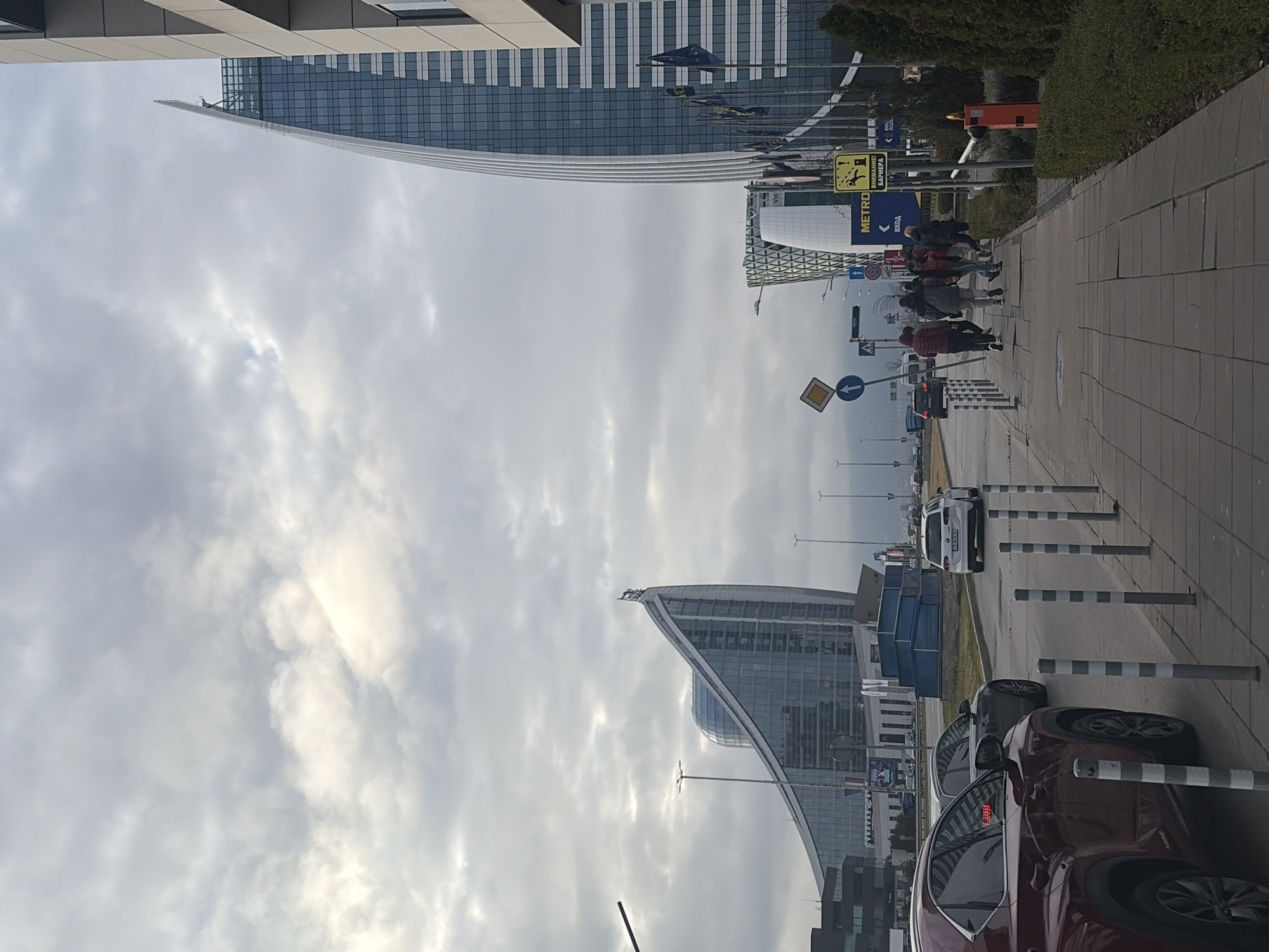 The image depicts an urban scene in Sofia, Bulgaria. Two prominent curved glass buildings rise into a cloudy sky. A group of people are walking along a paved sidewalk, adjacent to a street with parked cars and traffic. There are road signs and lampposts visible. The buildings appear modern and are located near a metro entrance, indicated by signage.