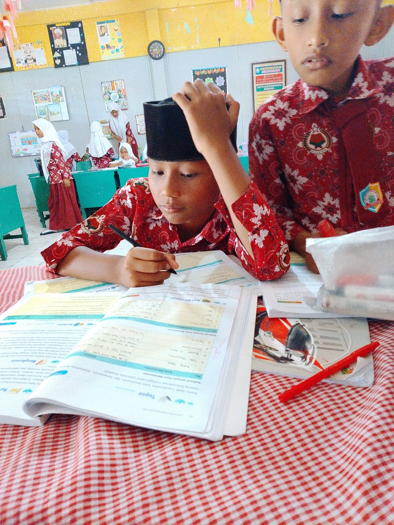 In a classroom environment, likely a primary school in Tembung, Indonesia, two young boys are the focus of the image. The boy in the foreground, wearing a red and white patterned shirt and a black peci (a brimless cylindrical hat), is seated at a table and appears to be writing in a textbook with a black pen. His hand is visible, holding the pen, and his gaze is directed downwards at the book. The other boy, partially visible in the background and to the right, is also wearing a similar red and white patterned shirt and is looking upwards. The classroom walls are decorated with educational posters, including a framed picture and what appears to be a clock. The table in the foreground is covered with a red and white checkered tablecloth and is laden with books and stationery. Other children, dressed in white hijabs and red and white patterned uniforms, are visible in the background, suggesting an active learning environment. The overall impression is one of a typical school day, with students engaged in academic activities.