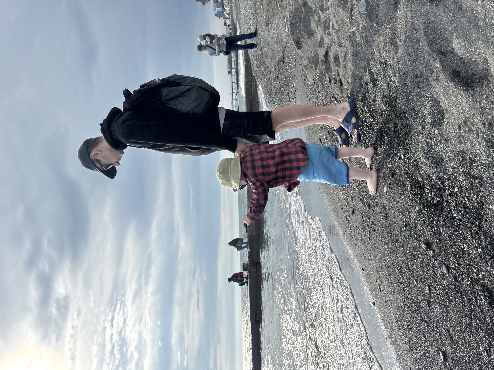 A father and son are standing at the edge of a beach in Sochi, Russia. The man is wearing a black hooded sweatshirt, black shorts, and blue sandals, and has a black backpack. The child is wearing a red and black plaid shirt, blue shorts, and a light-colored hat. They are barefoot. The man appears to be helping the child reach the water. In the background, there is a pier with people walking on it, and people are fishing on a concrete seawall. The sky is cloudy. The sand on the beach is dark gray.