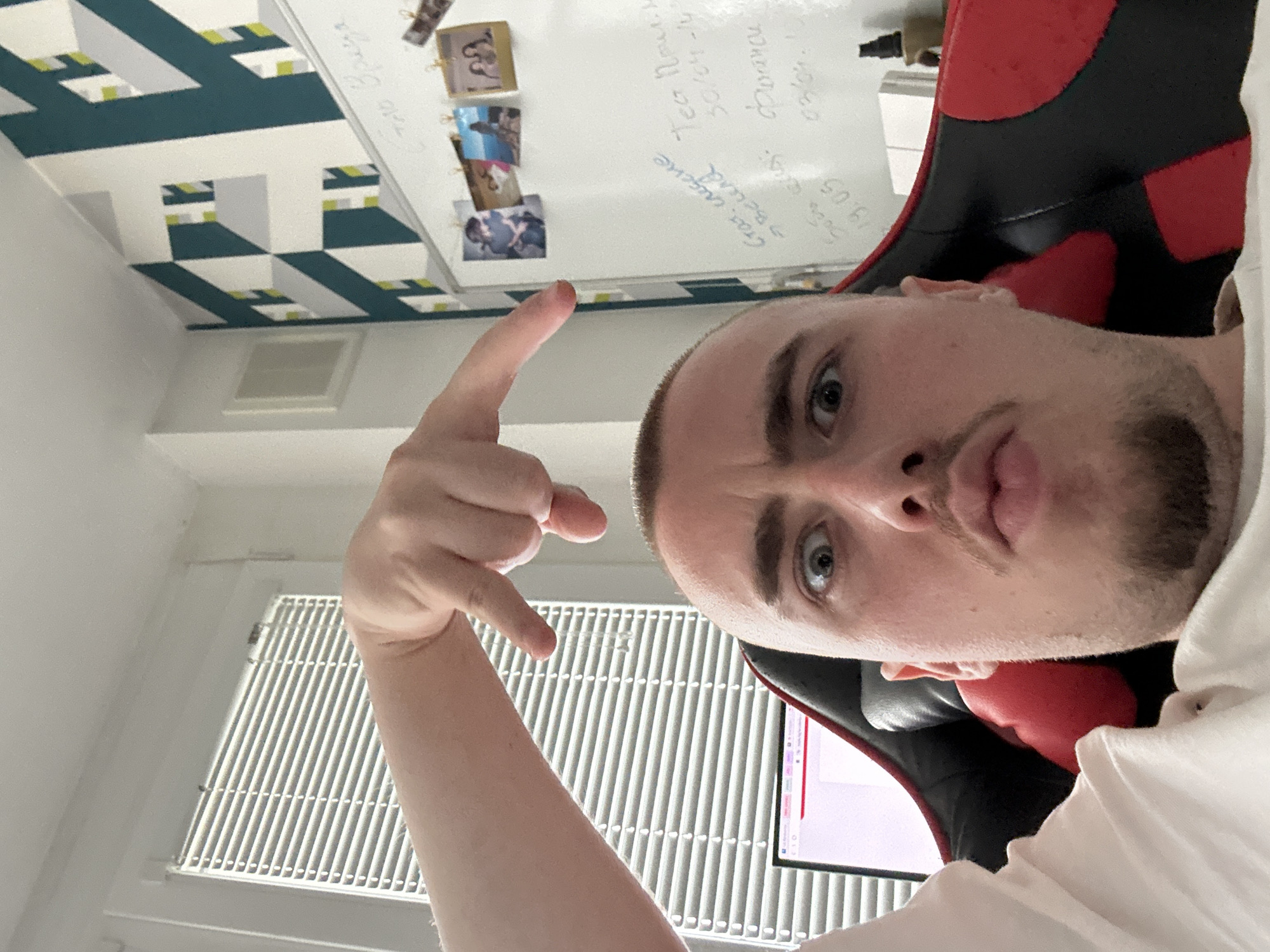 The image captures a young man in an indoor setting, likely a home office in Sofia, Bulgaria. He is seated in a prominent black and red gaming chair, looking directly at the camera with an intense, slightly puckered expression and wide blue eyes. He has a short beard and buzz-cut hair and is wearing a white t-shirt or hoodie. His right arm is raised, performing a gesture with his hand where his index finger points upwards and his thumb forms a right angle with the other curled fingers.
The background reveals a distinctive wall with a modern geometric pattern featuring teal, white, and lime green shapes, suggesting depth or a 3D effect. A white ventilation grate is visible on the upper wall. Above the man's head, a white board is mounted, adorned with several small, rectangular photographs clipped onto its surface. The whiteboard also features handwritten notes in Cyrillic. Visible text includes "С днём рождения" (Happy Birthday), "Тест Прошел" (Test Passed), "фотки" (photos), and dates such as "30/04-20" and "19.05". Below the whiteboard, a portion of a computer monitor with a red scroll bar is visible, suggesting a workspace. The image is taken from a low angle and is rotated 90 degrees clockwise.