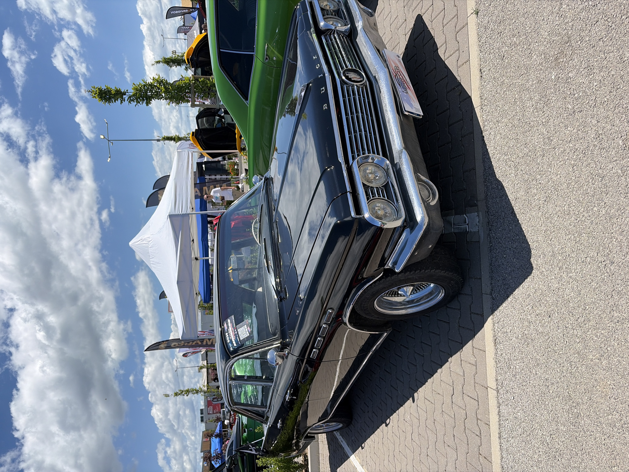 This is a medium shot, eye-level, outdoor photograph taken on a sunny day at what appears to be an outdoor event, possibly a car show, based on the presence of multiple vehicles, tents, and banners. The main subject is a shiny, black, vintage Buick sedan parked on a paved surface made of grey interlocking bricks. The car features chrome accents, including the grille, bumpers, trim, and side mirrors. The front of the car displays the Buick logo and the model name "BUICK" spelled out in individual letters on the hood. A license plate-like sign with red and blue markings and the words "MUSCLE CARS" is attached to the front bumper.
To the right of the Buick, a bright green vintage car is partially visible. Further in the background, there are white and blue tents, tall green trees, lampposts, and several colorful banners with writing on them. A few people are visible in the background, including one man wearing a white t-shirt standing near a tent, and another person standing in the open door of a yellow-hooded car. The sky is blue with fluffy white clouds.
The location is identified as Sofia, Bulgaria, specifically at the Sofia West Park, based on the information provided. The overall atmosphere suggests a community gathering centered around classic automobiles.