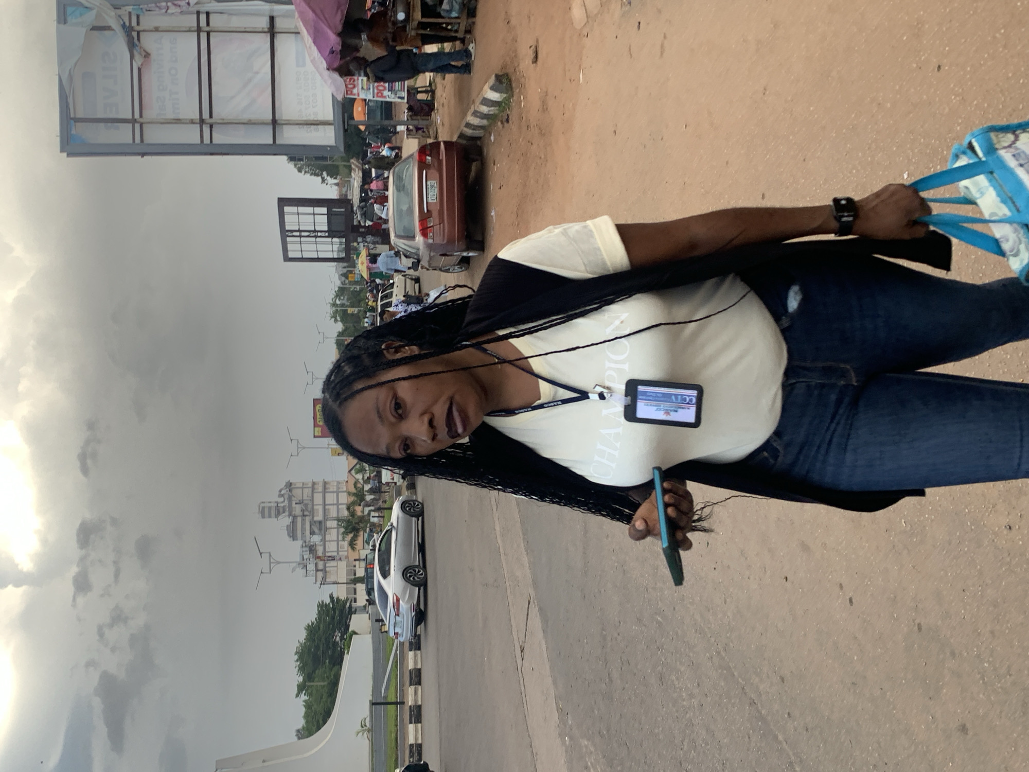 A person with dark braided hair stands in a street scene in Jos, Nigeria. They are wearing a light-colored t-shirt with "CHAMPION" visible in large letters, dark jeans, and a black jacket. A lanyard with a tag that reads "CCTV" is around their neck. In their right hand, they are holding a blue and white patterned bag, and in their left, a dark-colored smartphone.
The background shows a busy street with various vehicles, including a red car and a white car. Several people are visible, some walking and others gathered around stalls. A large billboard is present in the distance. The ground is a mix of dirt and pavement. The sky is overcast, suggesting cloudy weather. The lighting indicates it is daytime.