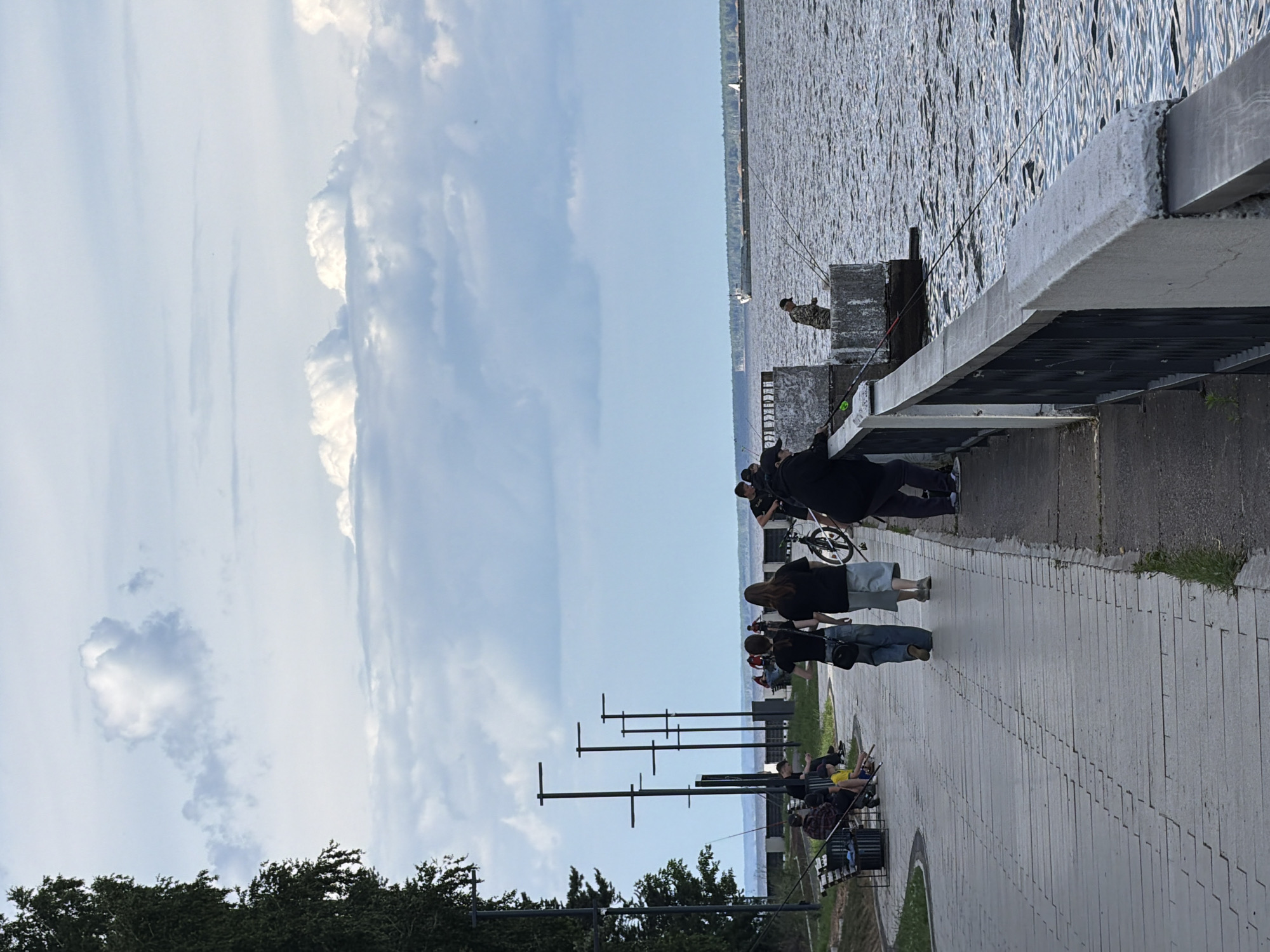 This is a vertical, eye-level shot of a waterfront promenade in Chaikovsky, Russia. The scene is bathed in the soft, diffused light of a partly cloudy day, suggesting it is likely morning or late afternoon. The sky is a pale blue, with large, white, puffy clouds scattered across it. The body of water to the right is a dark, rippling expanse, reflecting the light in a textured pattern.
Along the paved promenade, a group of people are seen engaged in various activities. In the foreground, several individuals are walking, with one person on a bicycle. Further down, more people are gathered, some appearing to be fishing from the elevated walkway. One person is leaning on a railing, looking out at the water, while others are seated on benches, also with fishing rods. The overall mood is relaxed and leisurely.
The architecture of the promenade includes concrete barriers and railings, with some metal posts lining the pathway. Trees with dark green foliage are visible on the far left of the frame. The image captures a common outdoor recreational scene by a large body of water. There is no visible text in the image.