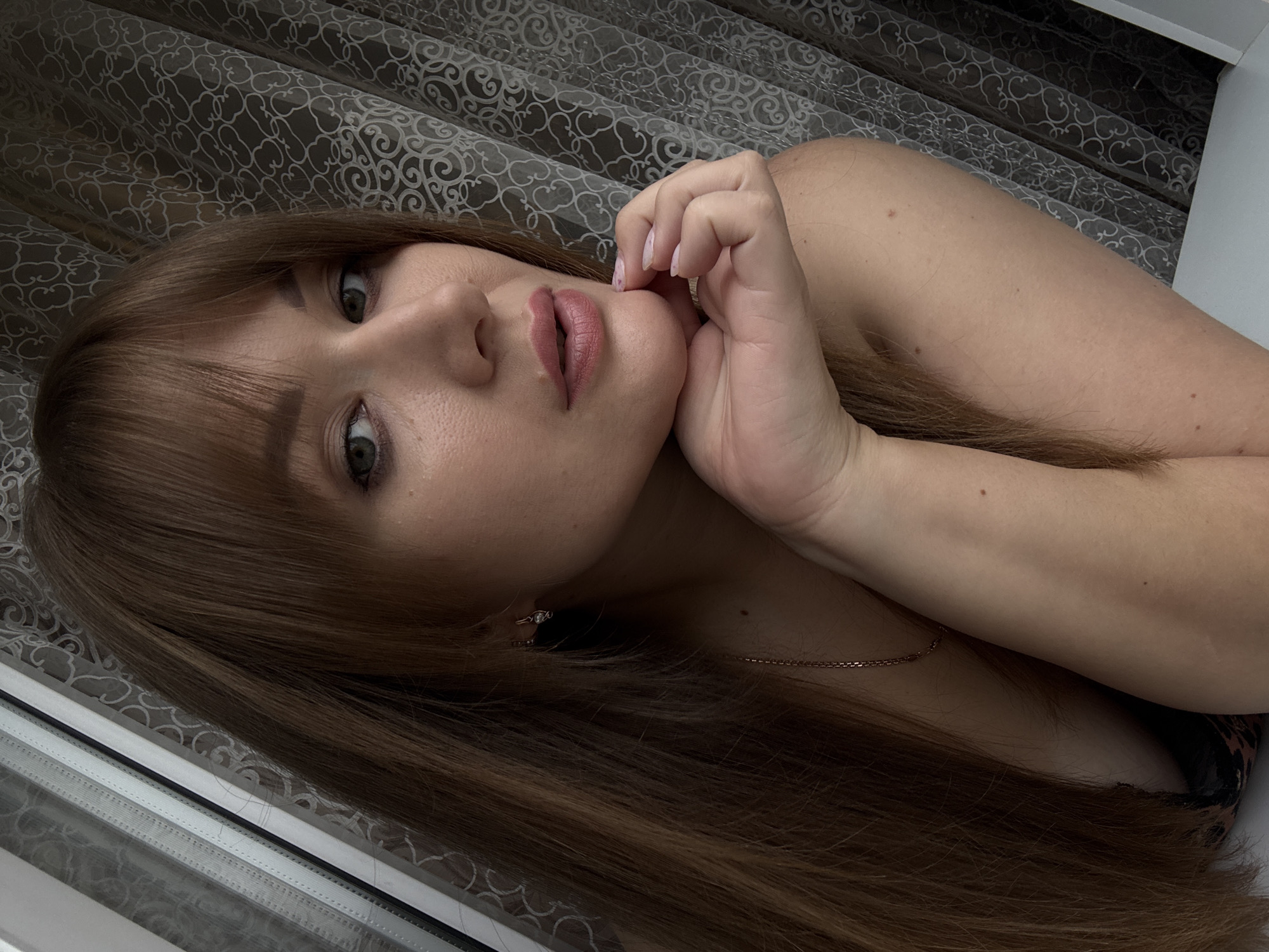 A young woman is lying down, with her head resting on her hand and her eyes looking into the camera. She has long, reddish-brown hair that falls around her shoulders and face. Her lips are painted a muted pink. She is wearing a delicate gold chain necklace and a ring on her finger. The background is a patterned curtain, and a window frame is visible on the left side of the image. The lighting suggests it might be daytime, but the mood is intimate and relaxed, possibly at home in Chaikovsky, Russia.