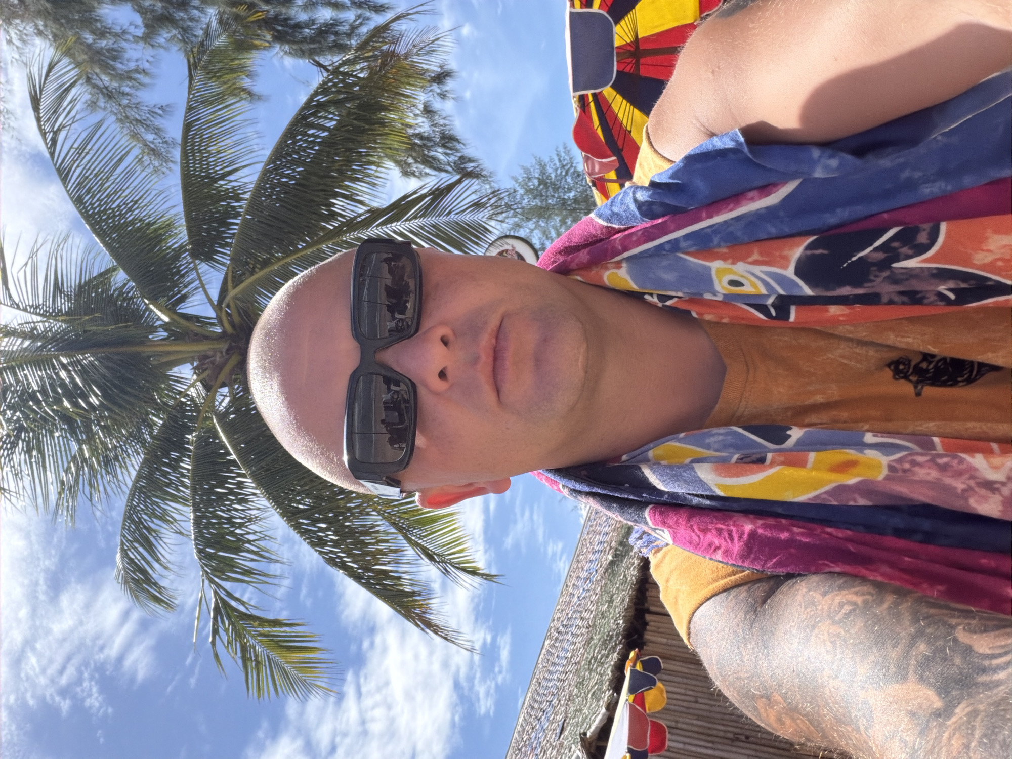 A bald man with sunglasses and a colorful towel around his neck is taking a selfie outdoors. He is wearing an orange t-shirt and has tattoos on his arm. Behind him, palm trees and a blue sky with clouds are visible. In the background, there is a thatched-roof structure and colorful umbrellas. The scene appears to be a beach or resort setting, possibly in the city of Choeng Thale, Thailand.