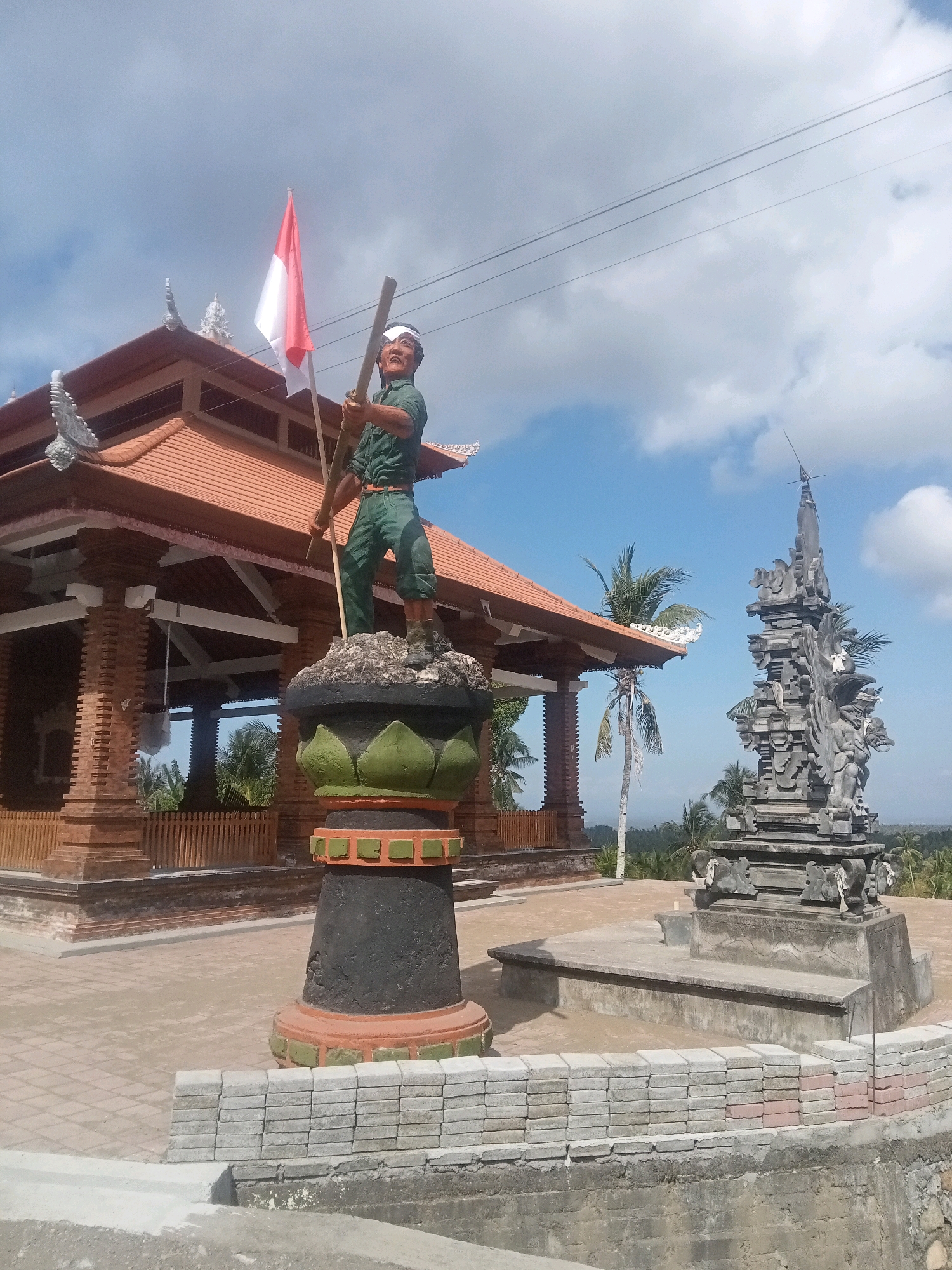 In Mendoyo, Bali, Indonesia, a statue of a man in a green uniform stands proudly, holding a wooden pole topped with the Indonesian flag. The statue is positioned on a decorative pedestal, with a traditional Balinese temple structure behind it. To the right, a carved stone monument adds to the cultural ambiance. The scene is set against a backdrop of palm trees and a bright, cloudy sky.