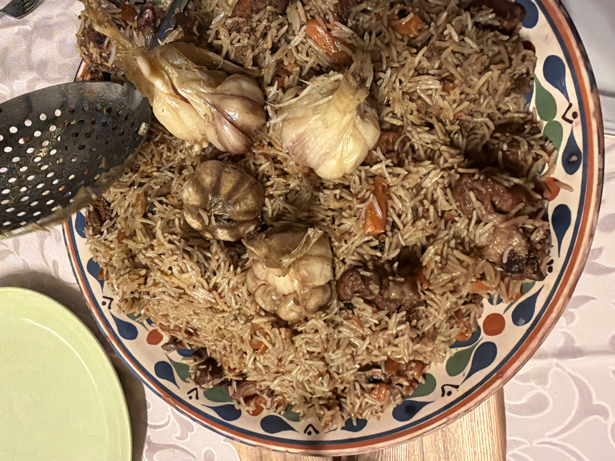 The image provides an overhead view of a large, round serving platter laden with a rich Plov, a traditional rice dish, set on a table. The Plov consists of long-grain rice mixed with pieces of meat, likely lamb or beef, and shredded orange carrots. Several whole, roasted garlic bulbs are prominently placed within the dish, appearing soft and caramelized.
A metal slotted spoon or skimmer is partially visible, resting within the Plov on the left side of the platter, suggesting it's ready for serving. The platter itself has a decorative rim featuring a colorful pattern of blue, orange, and green teardrop-like shapes and lines.
In the bottom left, a section of a plain, light green plate is visible. The table is covered with a white tablecloth that has a subtle, light-colored, ornate patterned design. A wooden surface is also partially visible beneath the tablecloth on the right.
The scene depicts an indoor setting, likely a dining area, where a meal is being served. The lighting appears to be artificial and warm, suggesting an evening meal. The dish's characteristics are consistent with Central Asian cuisine, which is common in Russia, aligning with the provided location context of Пролетарский, Russia. There are no people visible and no visible text in the image.