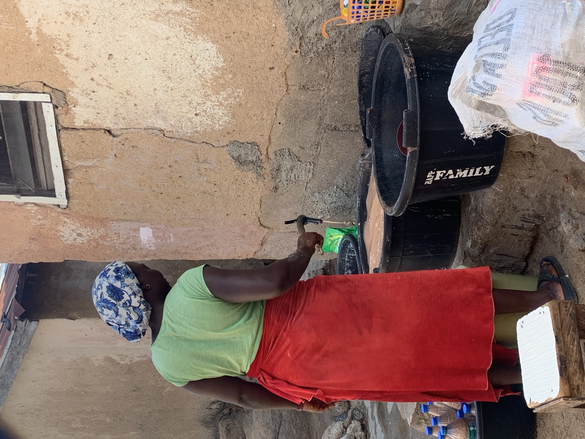 A medium shot depicts a woman in Unknown, Unknown, standing beside a wall while appearing to operate a water faucet. She wears a light green shirt, a red wrap-around skirt, and a blue and white head covering. Nearby are stacked black plastic buckets with the word "FAMILY" printed on the side. An opened box is on the ground. The wall in the background is light brown with weathered patches and a window. Other objects are visible in the surrounding environment.