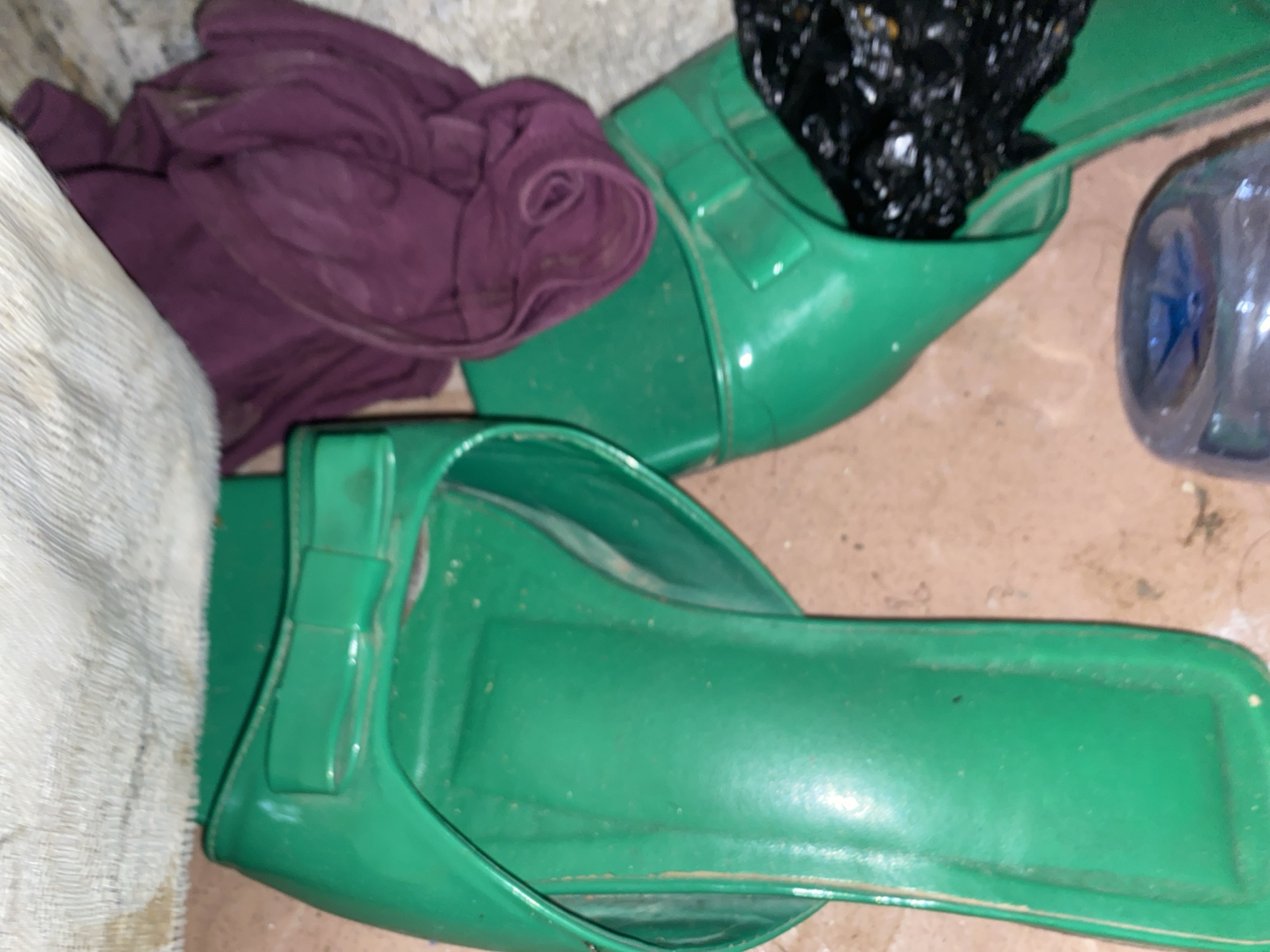 This media file shows a pair of bright green slide sandals placed on a dusty, light brown floor. One sandal is closer to the viewer and is fully visible, while the other is positioned behind it, partially obscured by a crumpled purple fabric and a black plastic bag filled with debris. A clear plastic bottle with a blue cap and some liquid inside is also visible to the right of the second sandal. The purple fabric appears to be a piece of clothing, possibly a skirt or a scarf, and it is bunched up next to the sandal. The black bag is overflowing with various dark-colored items. The scene is lit by an overhead light source, casting shadows and highlighting the textures of the floor and the objects. The overall impression is one of a cluttered, utilitarian space, possibly an outdoor storage area or a corner of a home. There is no visible text in the image. The setting appears to be in Jauro Yinu, Nigeria, suggesting a likely informal or outdoor environment. The time of day and weather are not discernible from the image.