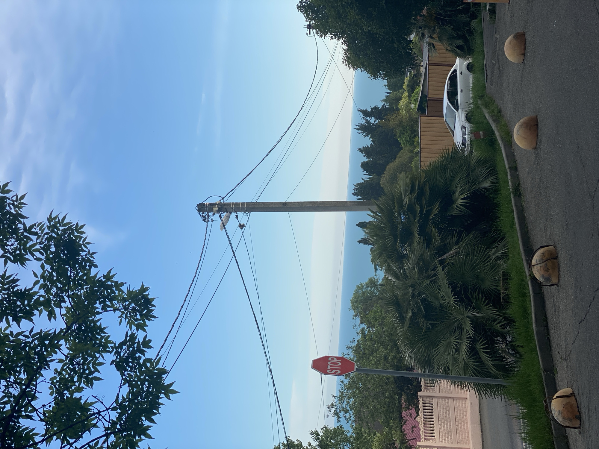 The image depicts a street scene in Sochi, Russia, with a view towards the sea. A concrete utility pole is visible with several power lines running overhead. A red stop sign is present on the right side of the road. The background reveals a coastal landscape with trees and buildings interspersed. A white car is parked alongside a light brown building with a darker brown roof. There are brown round stone bollards at the edge of the road.
