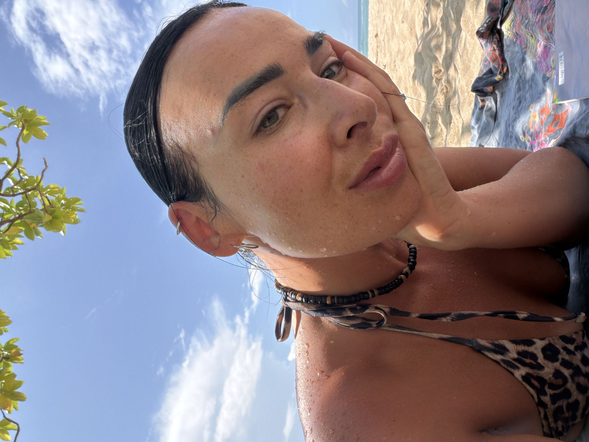 A woman is lying on a beach, possibly in Nusa Dua, Indonesia, based on the beach location and the presence of vegetation. She is wearing a leopard-print bikini and a beaded necklace. Her skin is damp. She is propped up on her left hand, with her face turned towards the camera. The background shows sand, a towel with colorful patterns, and a blue sky with sparse clouds and some green vegetation visible in the frame.