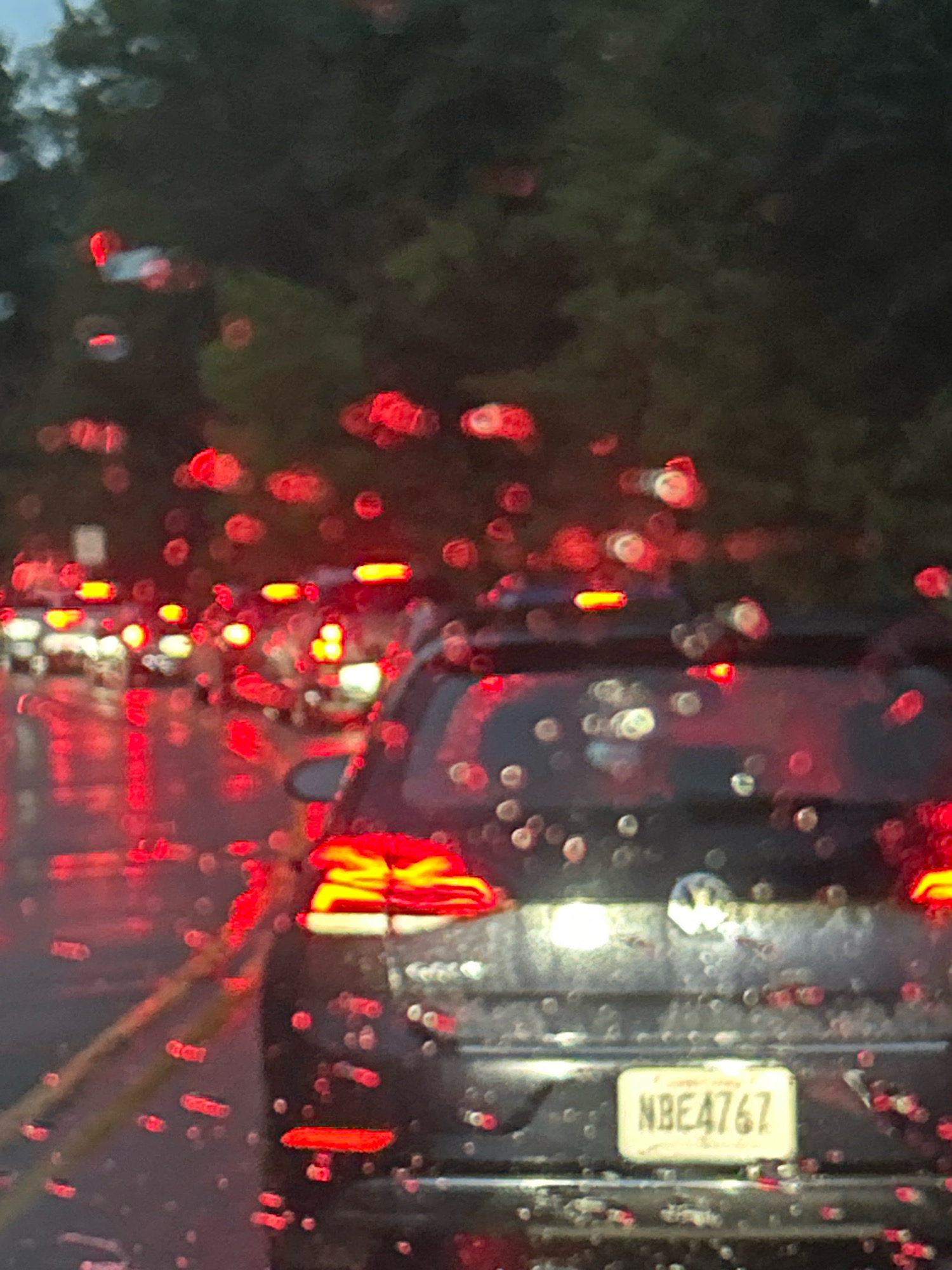The image shows a close-up view of the back of a dark-colored Volkswagen hatchback at night, with water droplets on the camera lens creating a blurry, bokeh effect. The car's license plate is visible and reads "NBE4767". The car's taillights are illuminated, as are the taillights of other vehicles in traffic ahead, indicated by red and white blurred lights. The background consists of dark trees and foliage, suggesting a road or highway setting within a city, likely Pittsburgh, United States of America, due to the context provided. The overall scene depicts a rainy night commute or traffic jam.