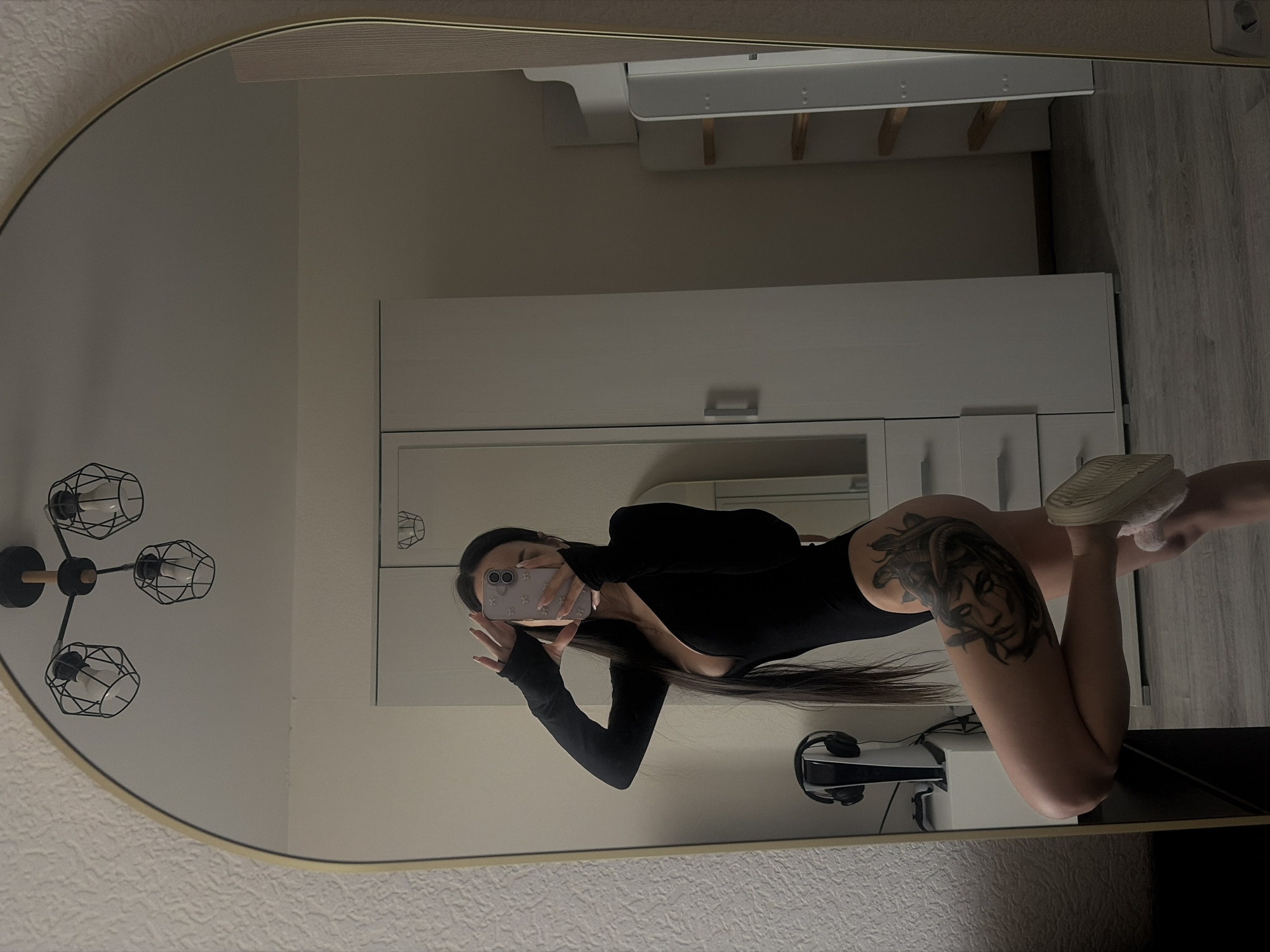 A woman is reflected in a mirror. She is wearing a black bodysuit and white sneakers. A tattoo of Medusa is visible on her thigh. The reflection includes a lamp, a white storage unit, a white shelf unit, a desk, and a PlayStation console. The environment appears to be a bedroom or apartment. The location is not discernible as Saint Petersburg, Russia, based solely on the image.