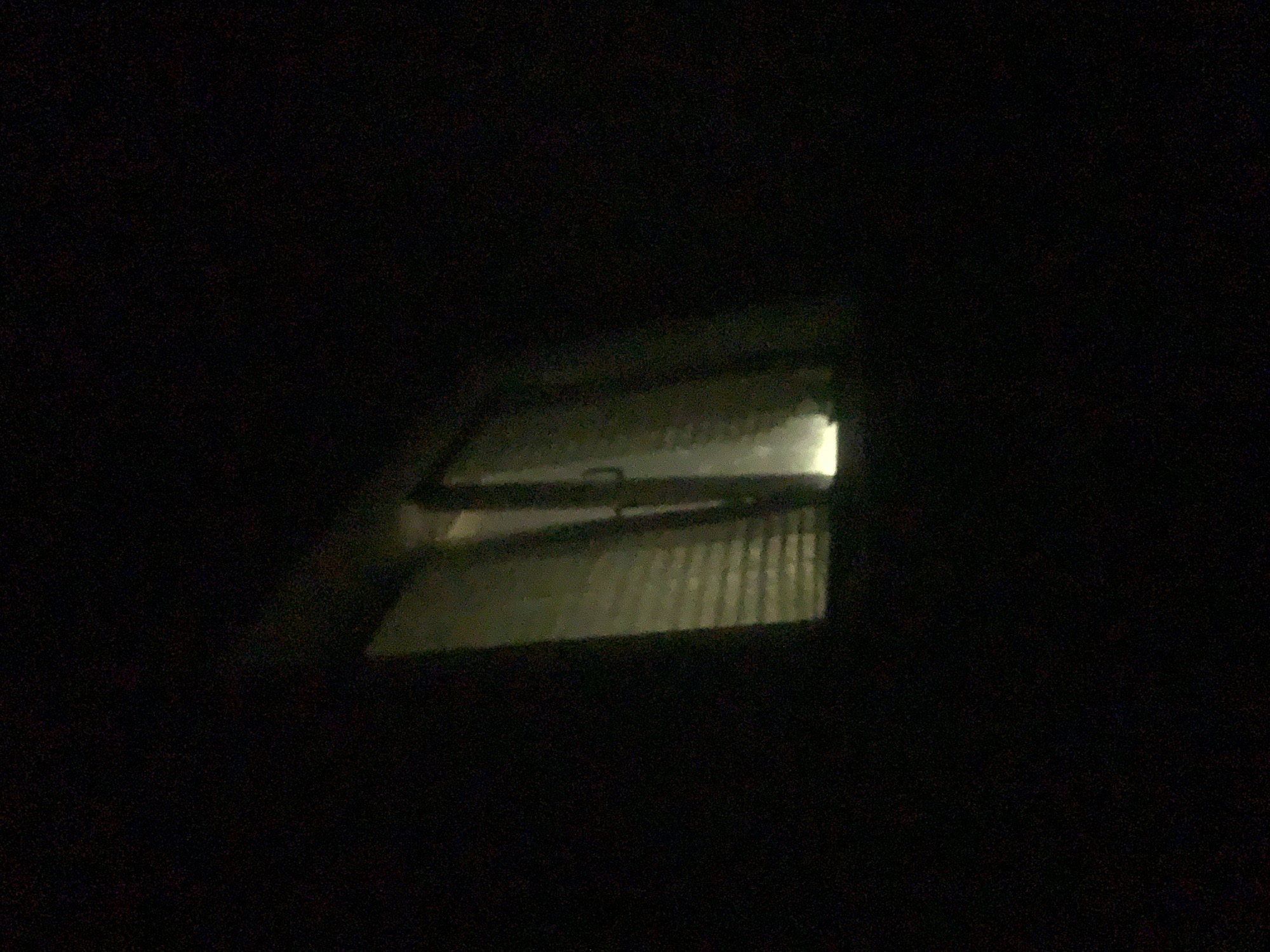 The image shows a dimly lit interior of a room, viewed through a window at night. The window appears to be a sliding glass door, with horizontal blinds visible behind it. Light emanates from inside the room, casting shadows and illuminating the blinds. The overall scene is dark and indistinct. There are no people, objects, or actions clearly discernible. The provided context that this scene is located in Minsk, Belarus, does not offer any visual cues within the image itself.
