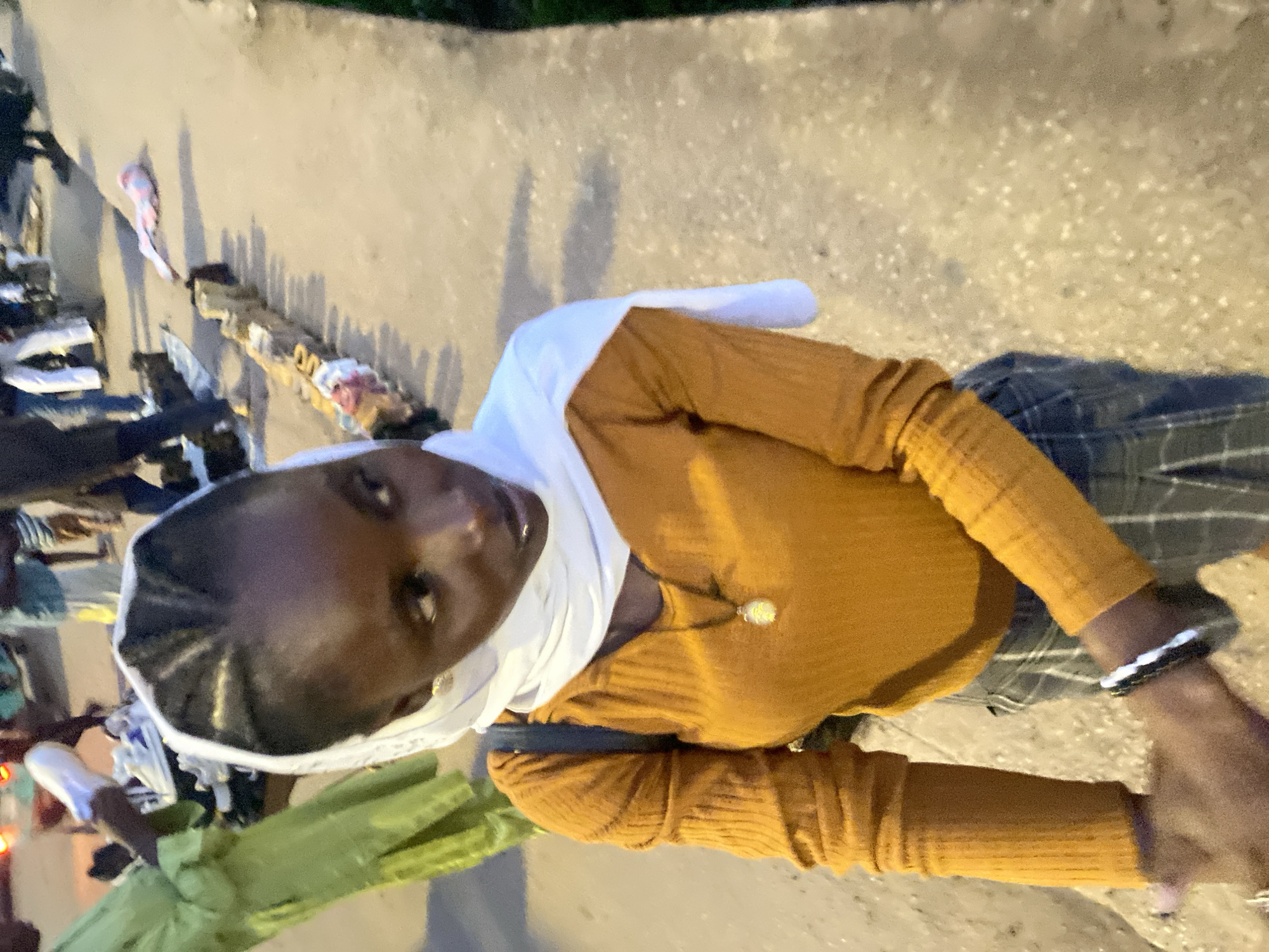 A young Black woman wearing a mustard-colored ribbed long-sleeved shirt and plaid pants is pictured outdoors in what appears to be a market setting in Bukuru, Nigeria. She has a white headscarf wrapped around her head and neck, and a necklace with a light-colored pendant. Her hair is braided in thin cornrows. She is looking to her right, with her gaze directed away from the camera. A dark backpack is visible on her back. Her right hand, adorned with bracelets, is extended forward. In the background, several other people are visible, along with a display of goods, possibly shoes and clothing, laid out on the ground. The ground is paved with concrete. The lighting suggests it is late afternoon or early evening.