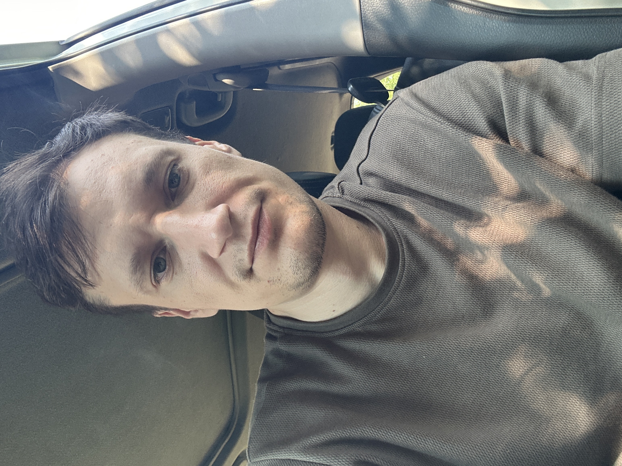 A close-up, low-angle selfie taken from inside a vehicle shows a light-skinned man with dark hair, a fair complexion, and light blue eyes. He is wearing a dark, textured shirt. The man is looking directly at the camera with a neutral expression. The interior of the car is visible, including the headliner and the edge of a seat. Sunlight filters in, casting dappled shadows across the man's face and shirt. The background is out of focus, but suggests an outdoor environment. No location in Shatava, Ukraine is identifiable in this image.